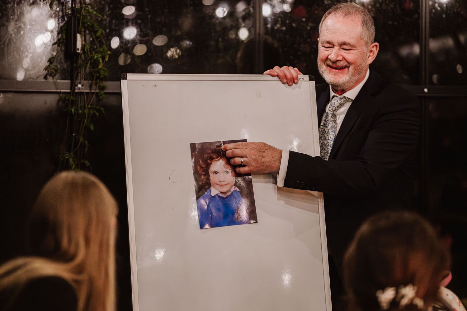 Smiling man in suit presenting a childhood photo on a large white board at an indoor event. The Cornish Place, Wedding