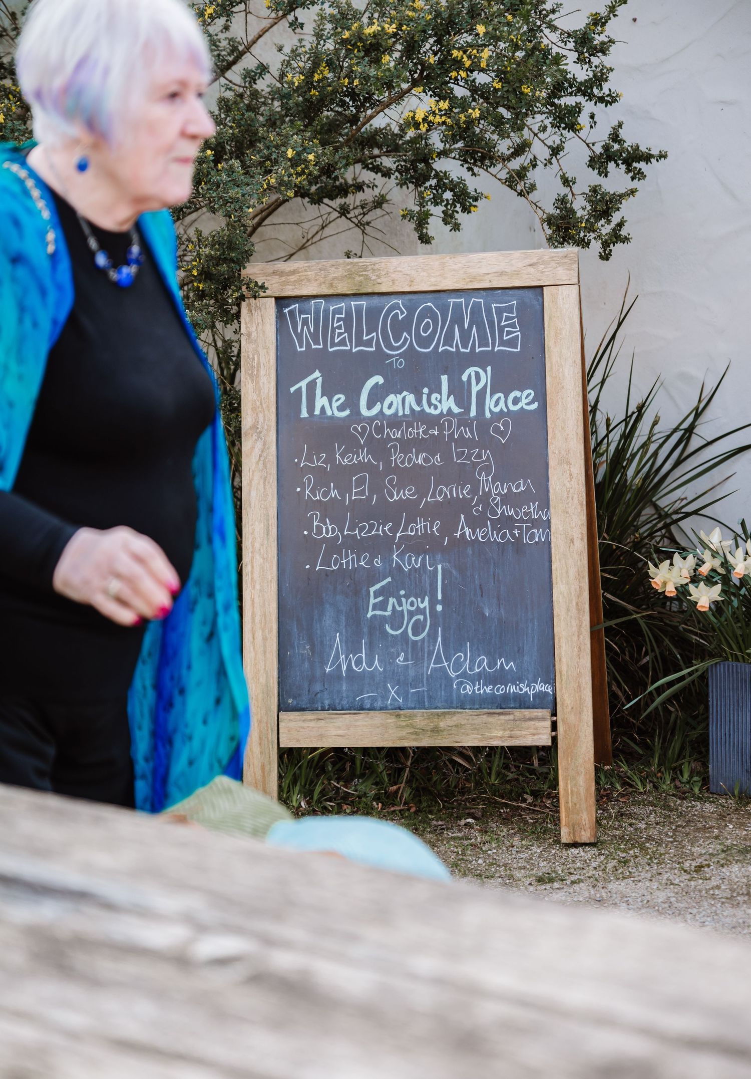 Elderly woman in blue jacket stands beside a chalkboard welcome sign for The Cornish Place outdoor event.