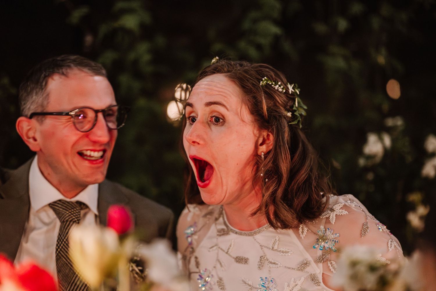 Bride with flower crown reacts with surprise and joy beside groom in glasses and tie at wedding reception.