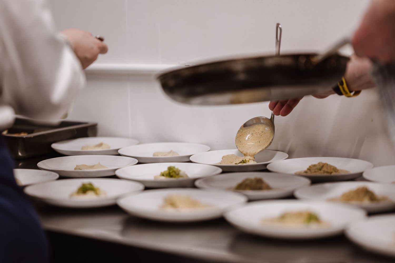 Chefs plating gourmet dishes, ladling creamy sauce onto white plates in a professional restaurant kitchen.