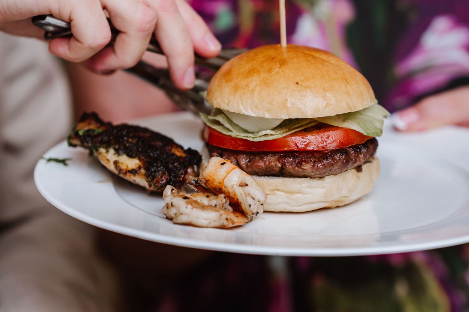 A juicy burger with tomato and greens on a white plate, served with grilled meat skewers.