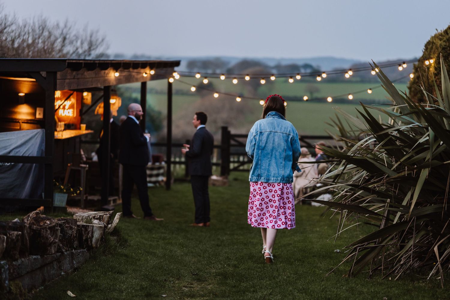 Guests mingle outdoors at dusk near a rustic bar with string lights at a countryside wedding venue. The Cornish Place, Cornwall wedding