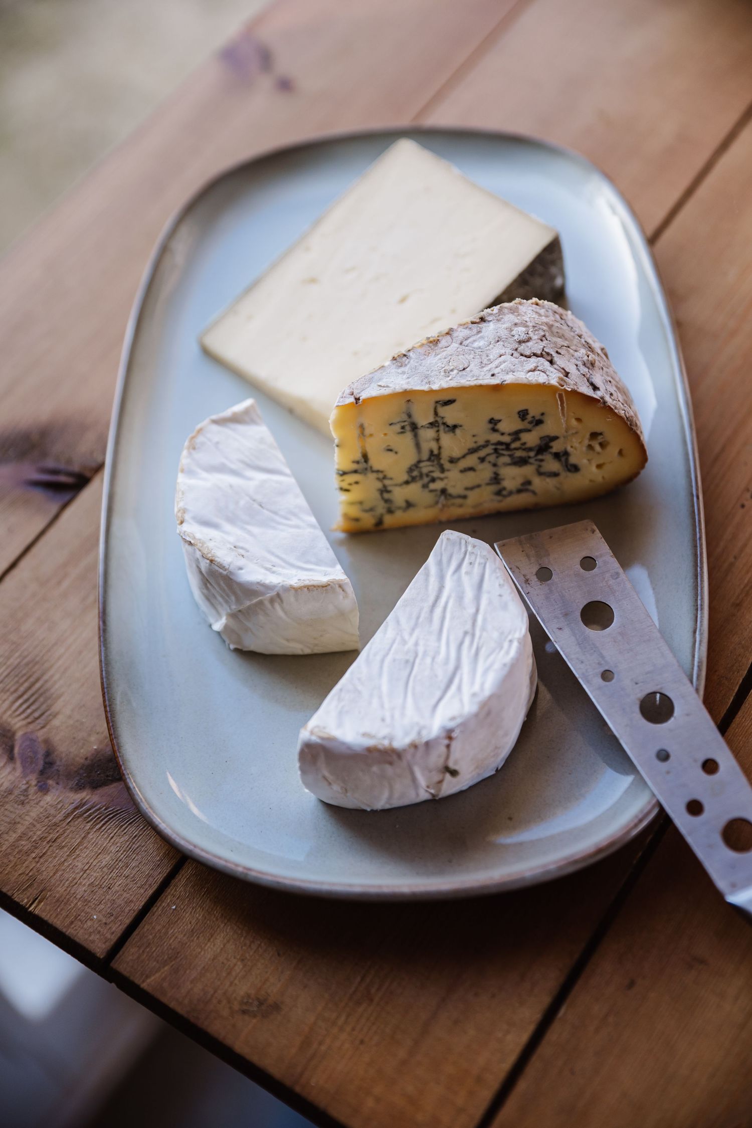 A rustic cheese board featuring brie, blue cheese, and aged varieties on a gray ceramic plate with a knife on a wooden table.