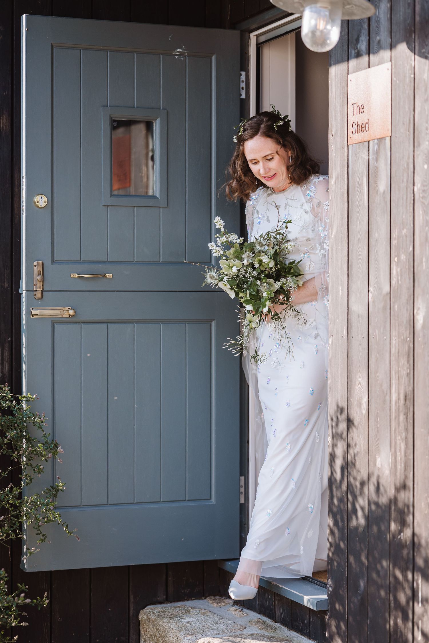 Bride in white embellished gown holding wildflower bouquet, standing at a blue stable door entrance.