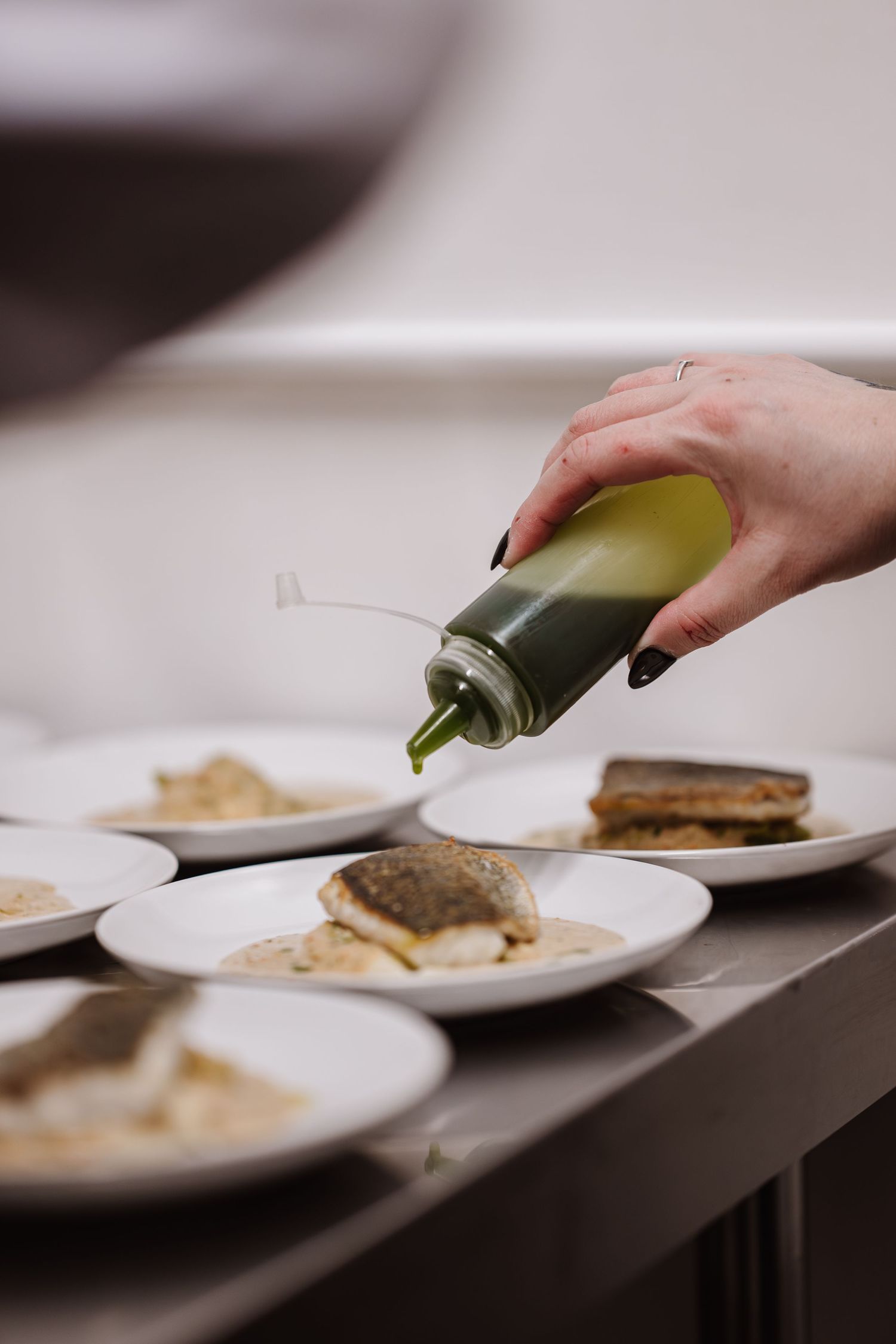 Chef drizzling green herb oil from squeeze bottle onto plated fish dishes in professional restaurant kitchen.