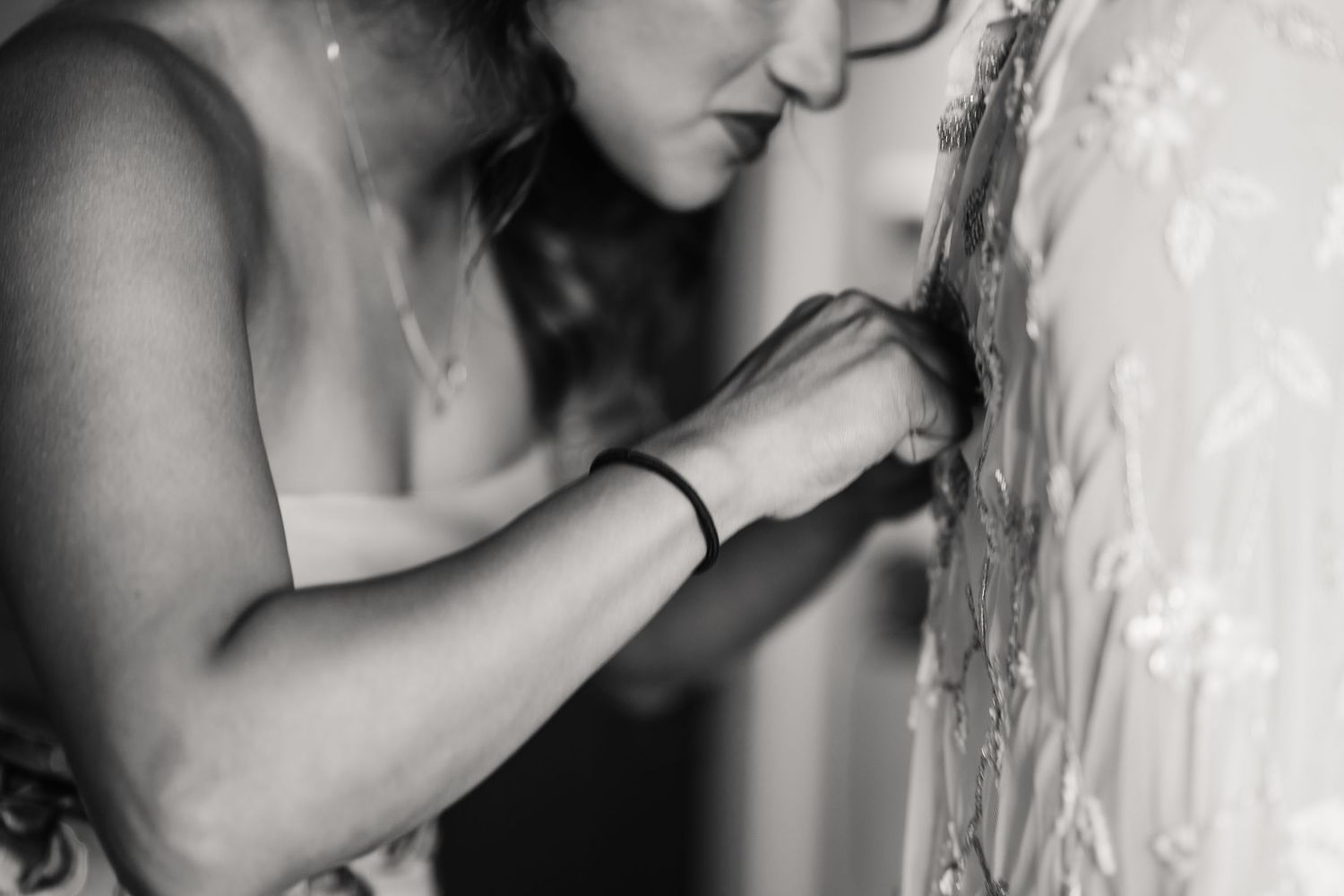 Black and white photo of a woman carefully fastening buttons on a lace wedding dress at The Cornish Place