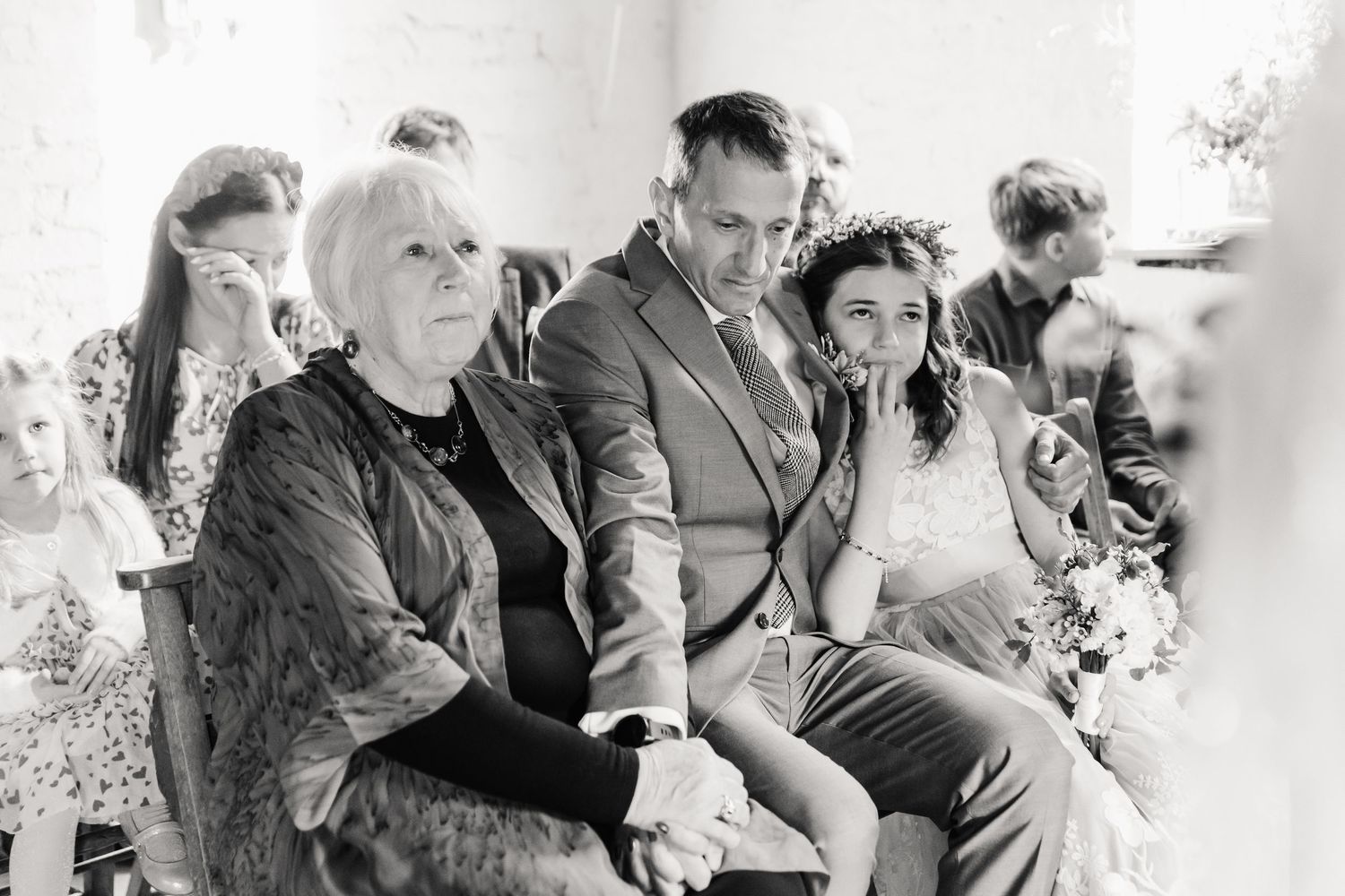 Black and white wedding photo of Mother of the groom and groom seated with family during ceremony. The Cornish Place, Wood shed wedding