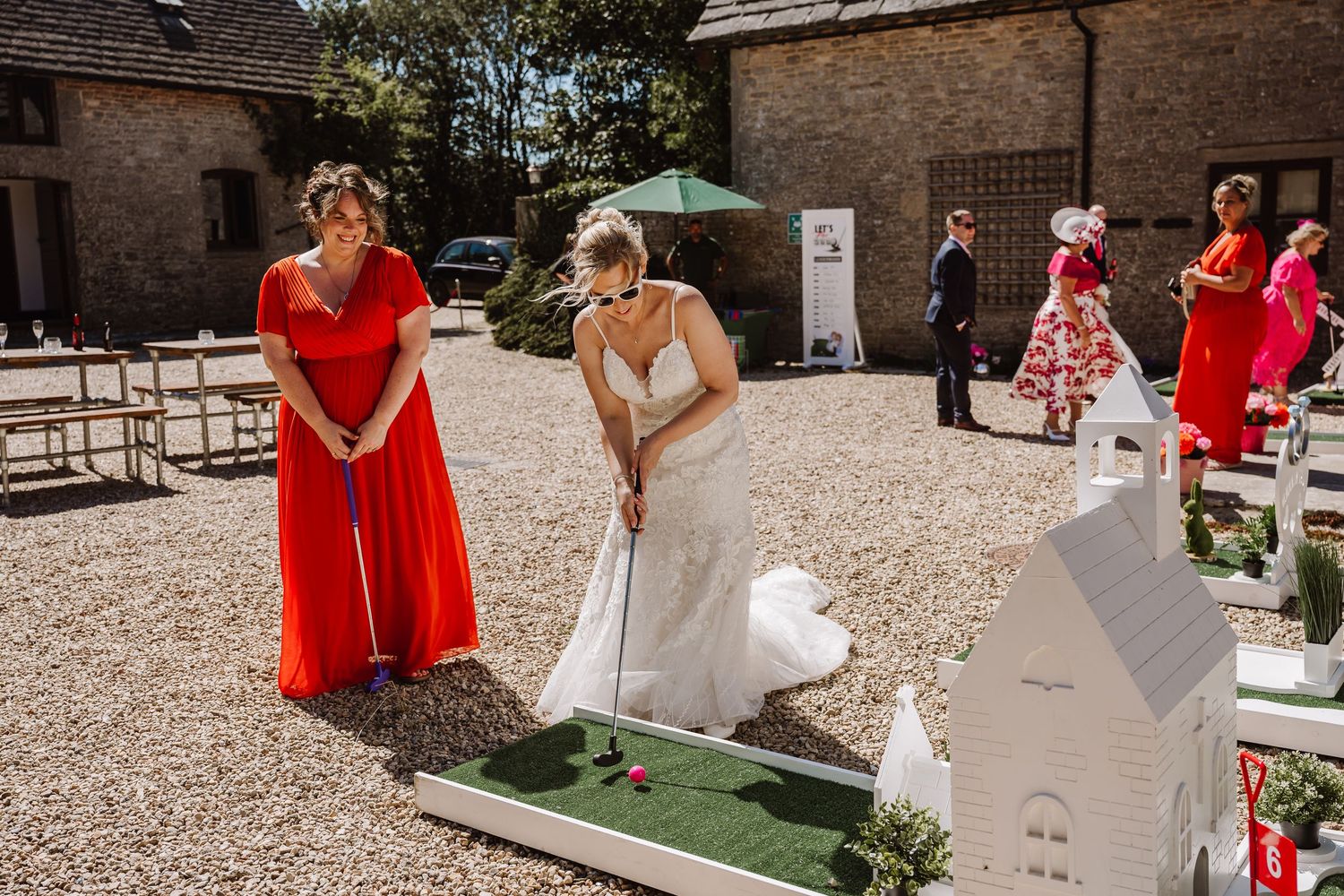 Bride in white gown plays mini golf at outdoor wedding reception while bridesmaid in red dress watches at Kingston Country Court Yard
