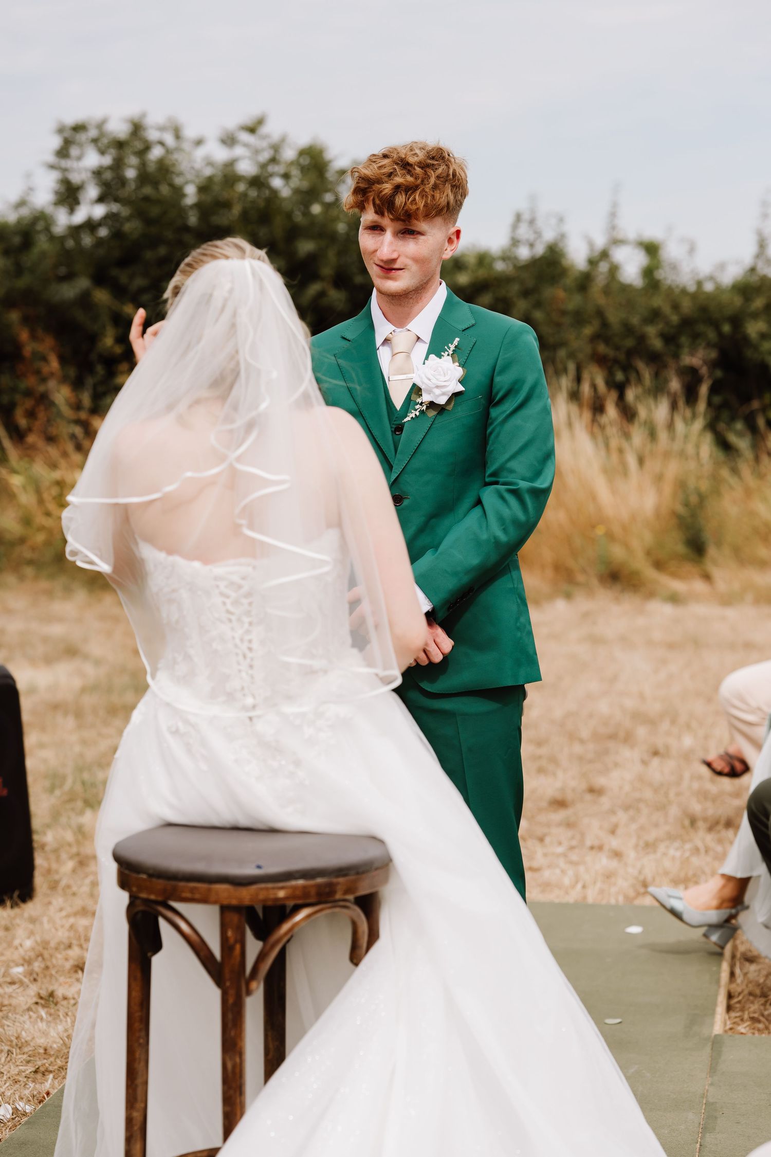 Groom in green suit smiles at bride in white gown and veil during outdoor wedding ceremony at Moor Farm, Weymouth Dorset