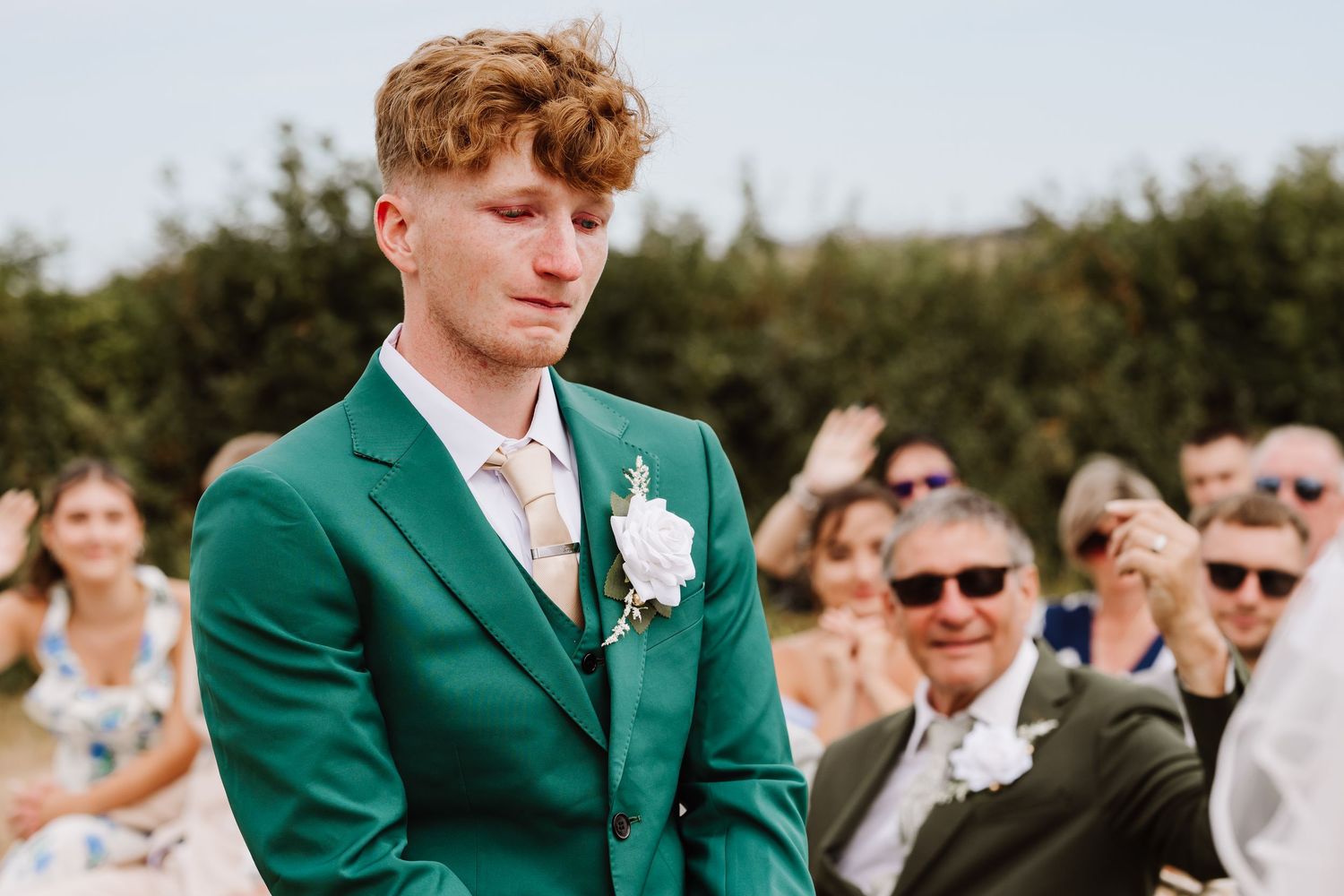 Emotional groom in green suit with boutonniere reacts as bride walks down the aisle at Moor Farm, Weymouth Dorset outdoor wedding.