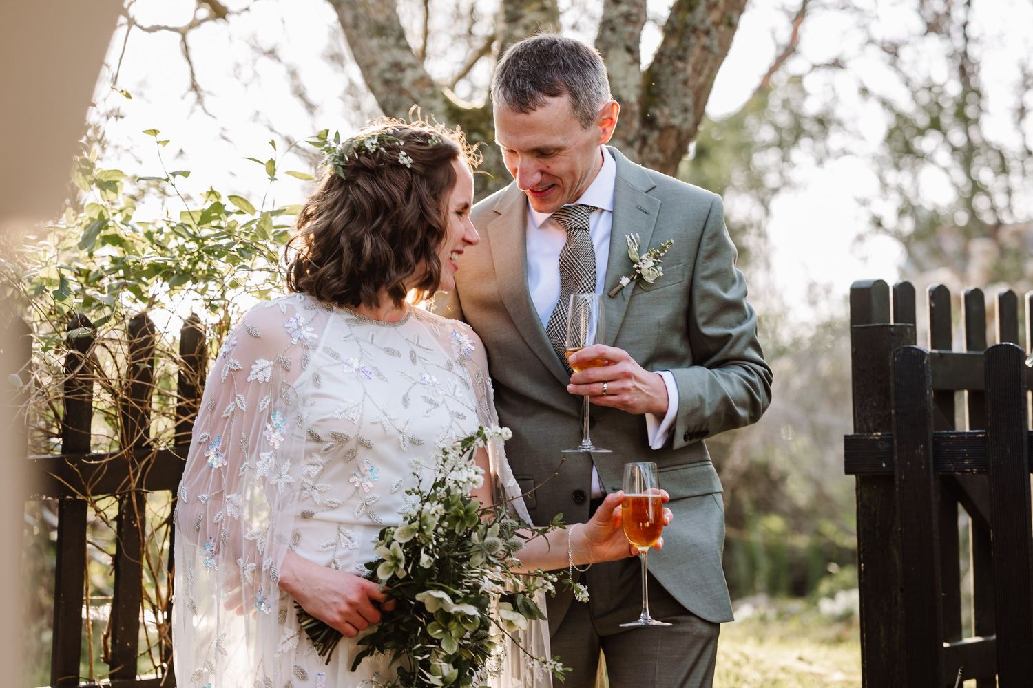 Bride in lace gown holding bouquet stands with groom in gray suit holding champagne glass outdoors at wedding at The Cornish Place wedding