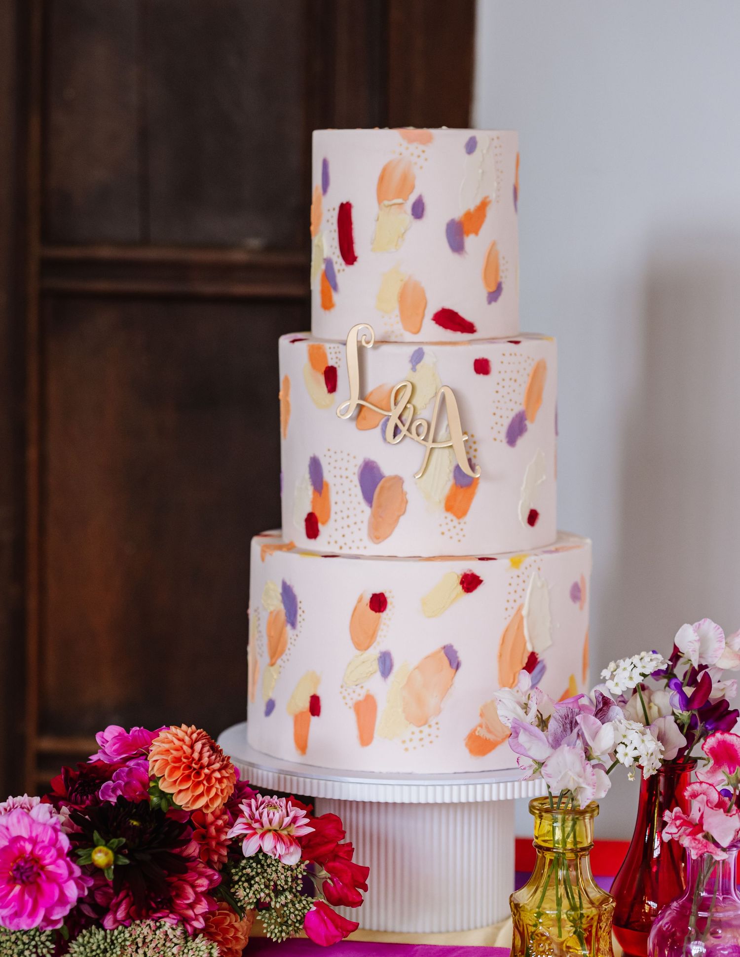 White wedding cake decorated with peach and red brushstrokes displayed among colorful flowers on a dessert table.