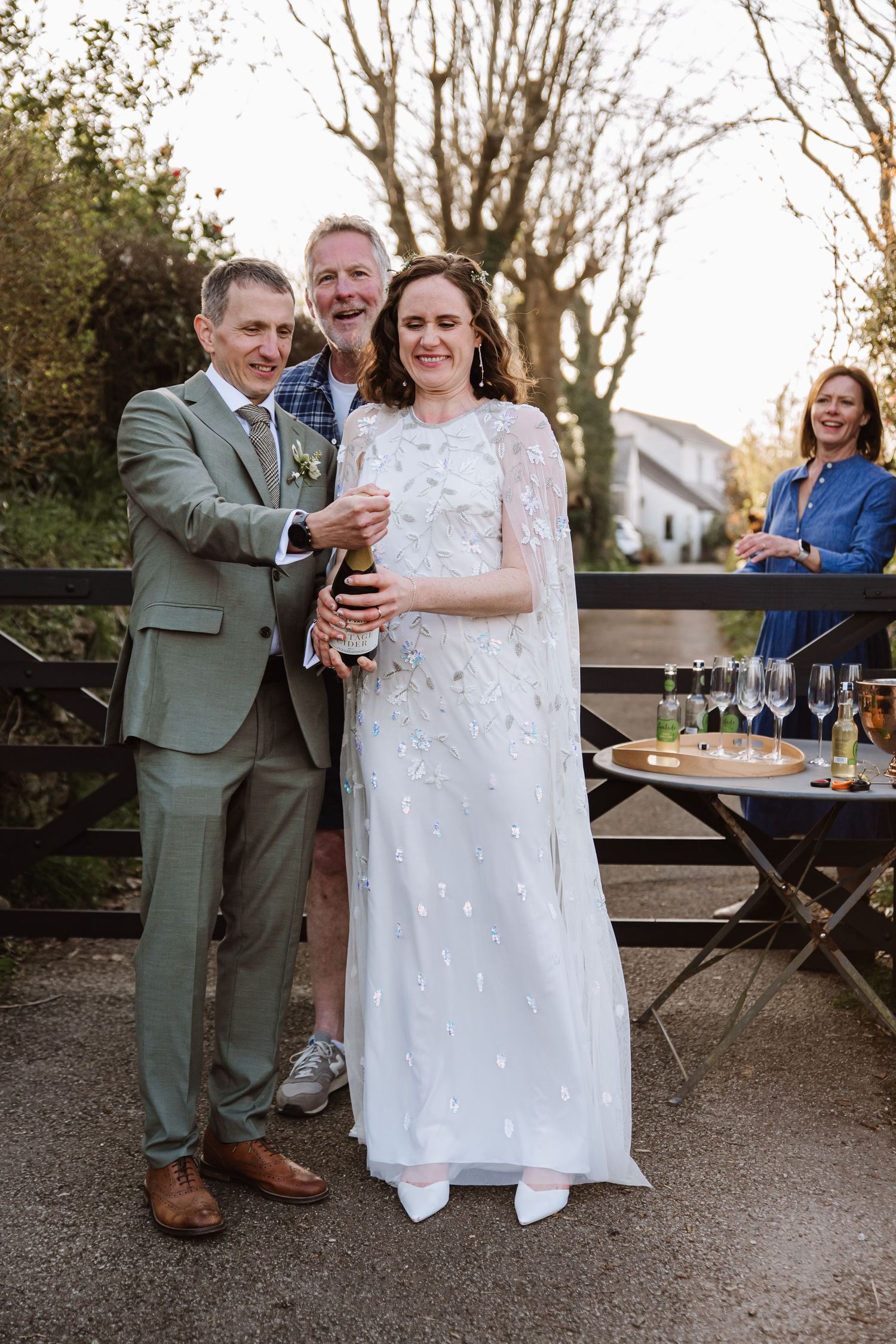 Bride and groom popping champagne bottle outdoors at wedding celebration with guests smiling in background. The Cornish Place, Wood shed wed