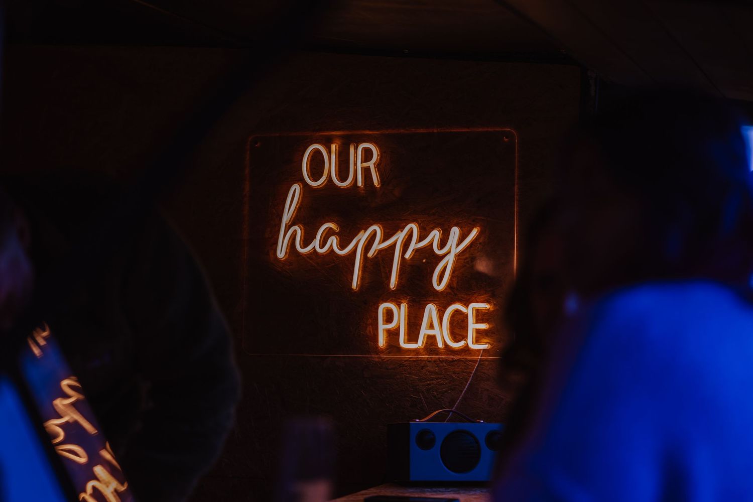 Glowing orange neon sign reading Our Happy Place mounted on a dark wall in a dimly lit venue. Outside bar at The Cornish Place, wedding venu