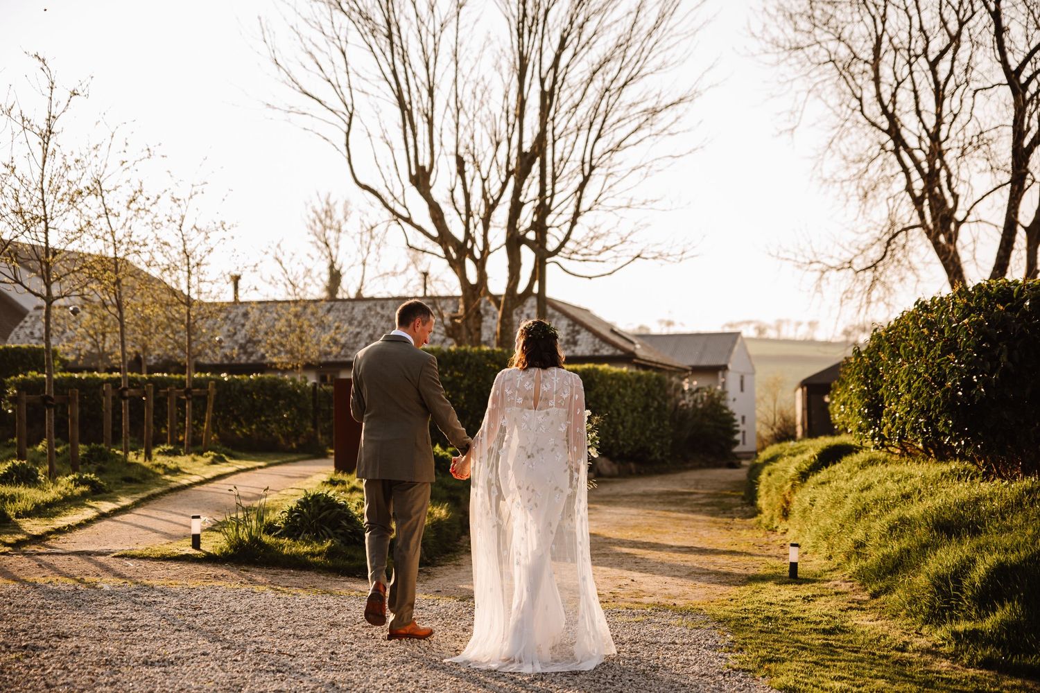 Bride and groom walking hand in hand through a rustic venue garden at golden hour on their wedding day. The Cornish Place, Wood shed wedding