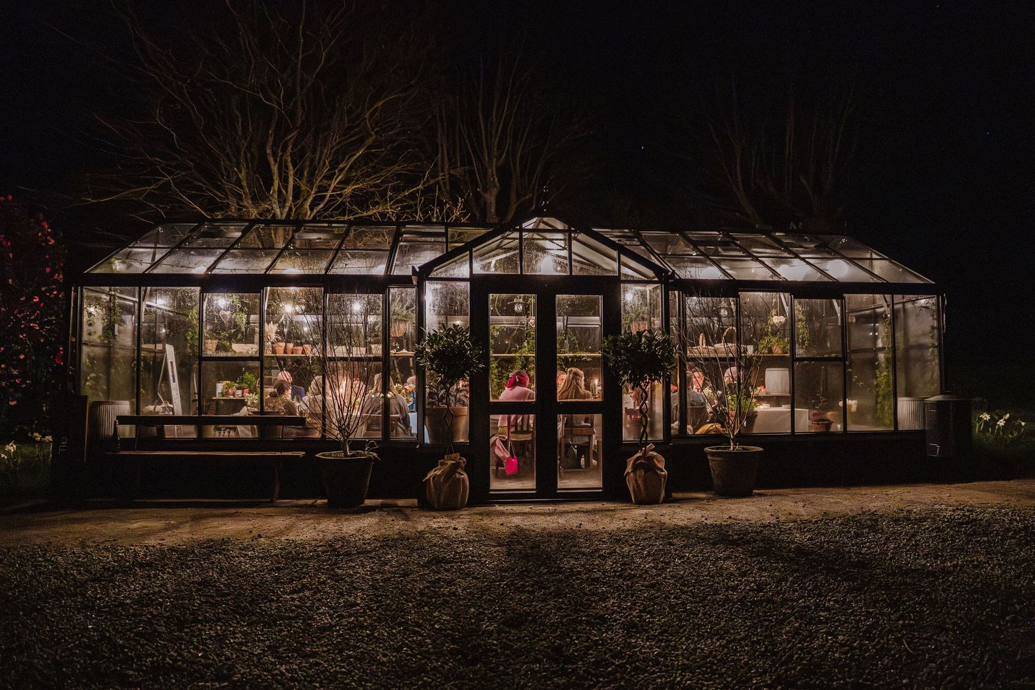 A warmly lit glass greenhouse glows at night, filled with plants and fairy lights, creating a magical garden display.
