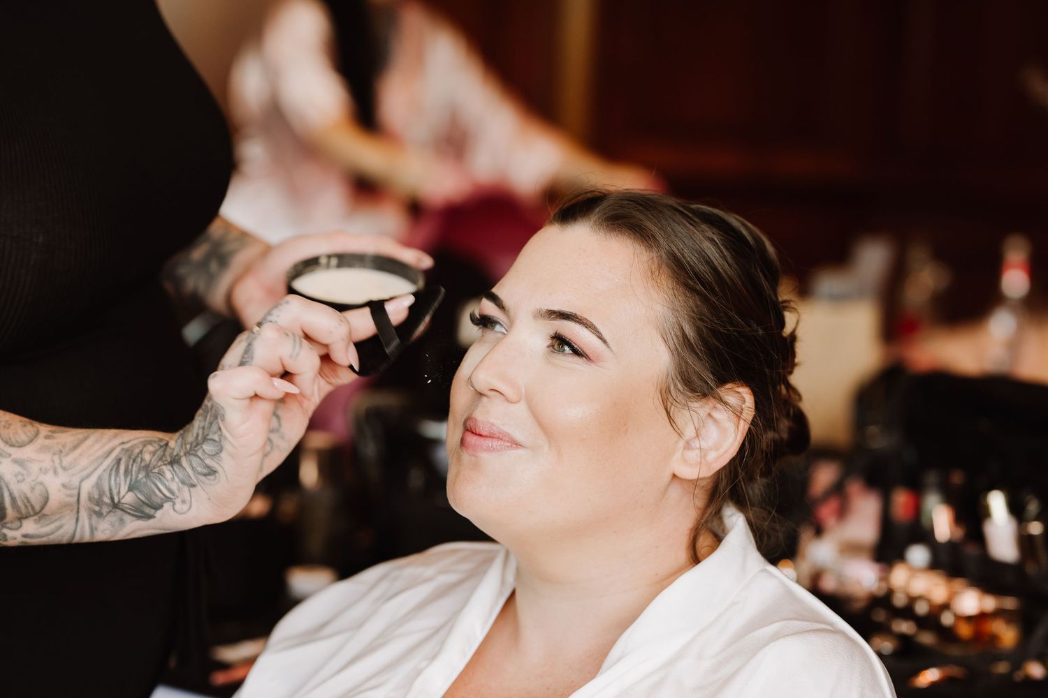 Bride in white robe smiling as makeup artist with tattooed arm applies makeup before wedding ceremony at Southampton wedding