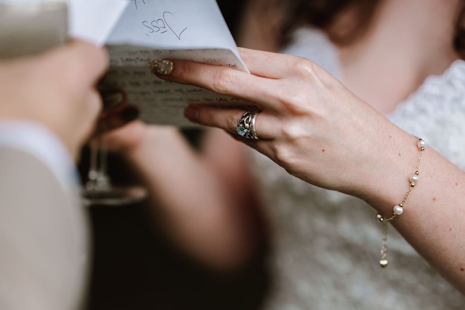 Close-up of bride's hand holding wedding vows letter, wearing elegant pearl bracelet and vintage engagement ring at Symmondbury Tithe Barn,