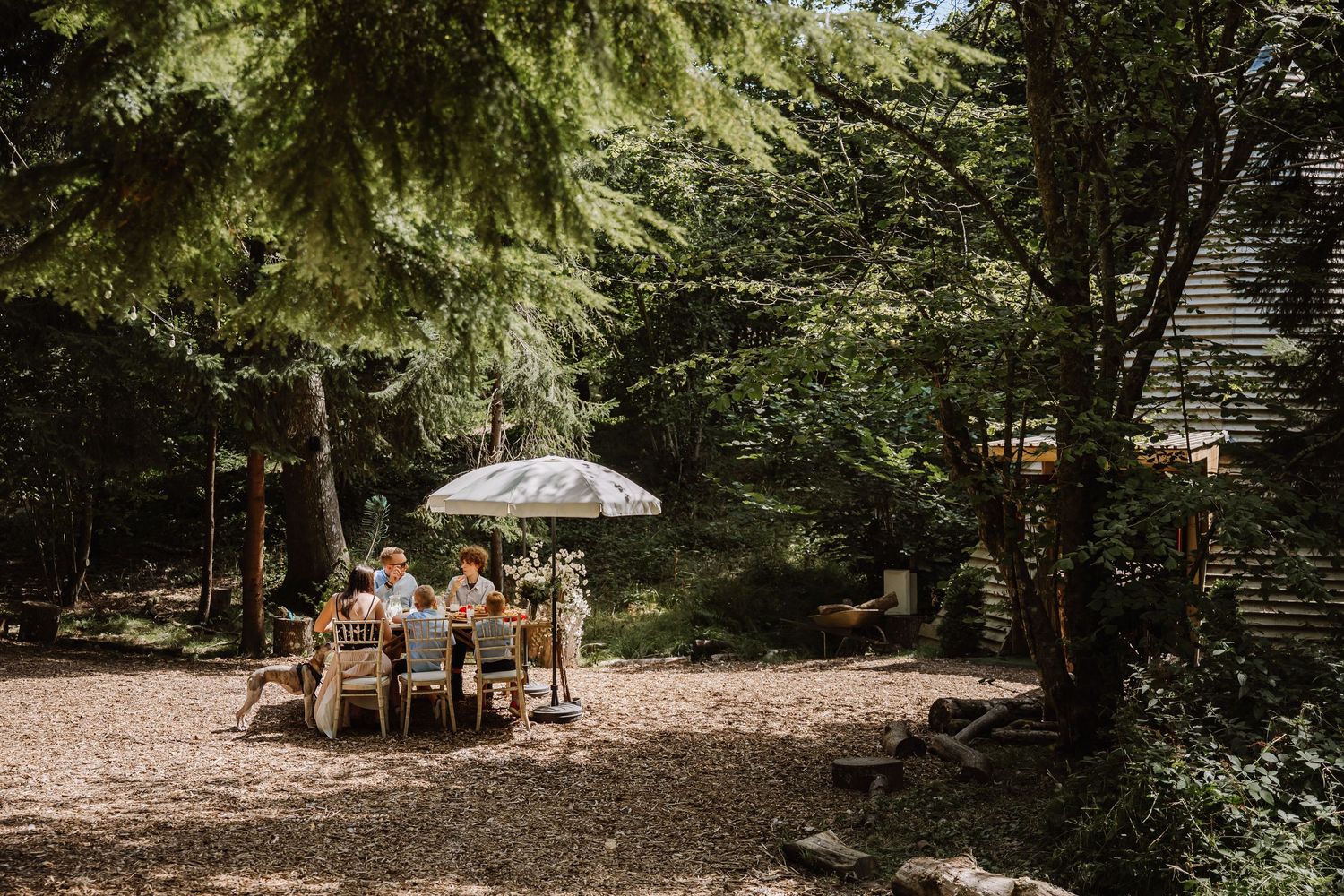 Moonacre Elopement Wedding, Dorset dining outdoors under a white umbrella surrounded by lush green trees in a serene woodland setting.
