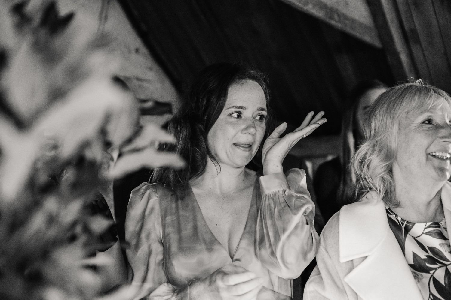 Black and white photo of a woman gesturing expressively at a wedding reception, laughing with guests nearby.
