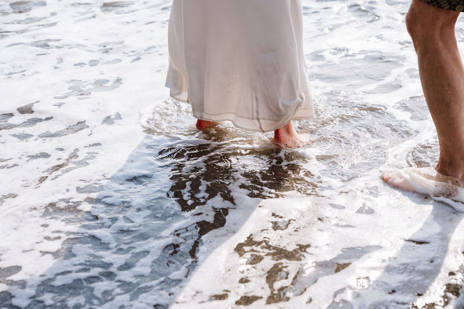 Two people standing barefoot in ocean waves, one wearing a flowing white dress on a sandy beach Cornwall beach wedding day