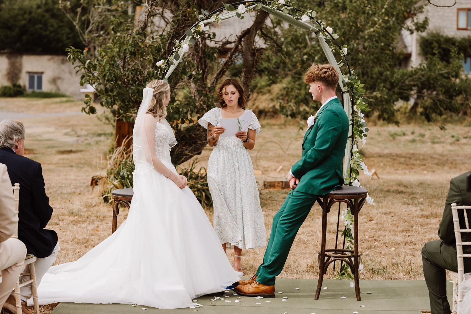 Bride and groom exchange vows outdoors under floral arch, officiant in white dress, groom wearing green suit at Moor Farm, Weymouth Dorset.