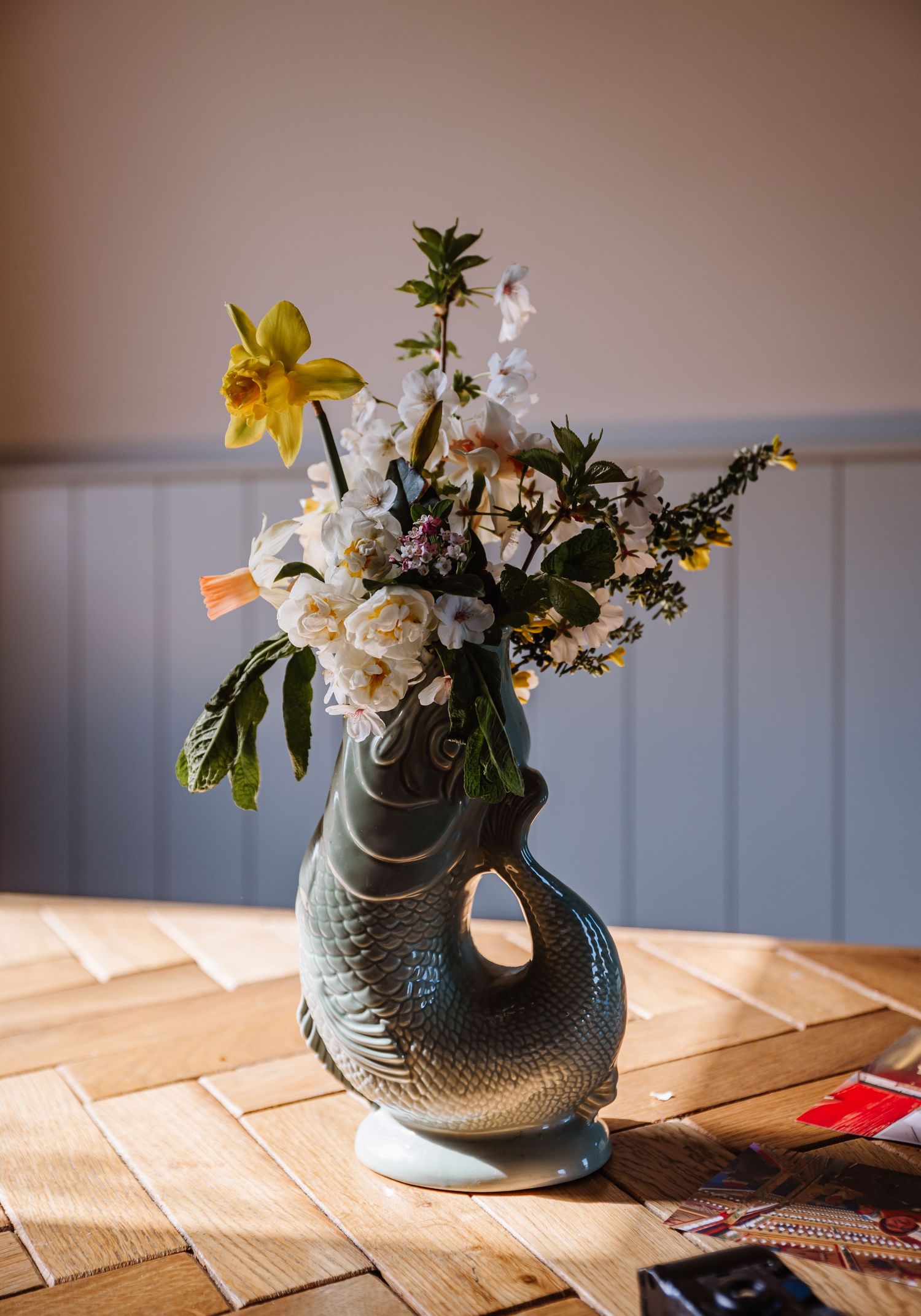 A fishshaped ceramic vase holding spring flowers including daffodils and white blooms on a wooden table. The Cornish Place, Cornwall wedding
