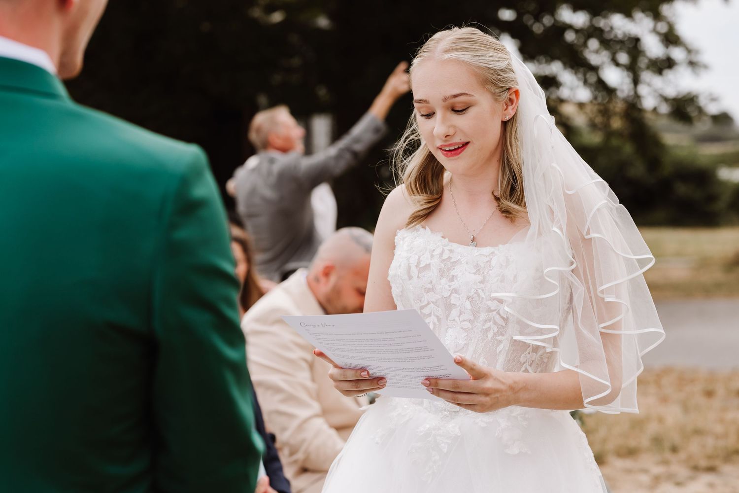 Bride in white lace gown and veil reads wedding vows from paper during outdoor ceremony at Moor Farm, Weymouth Dorset