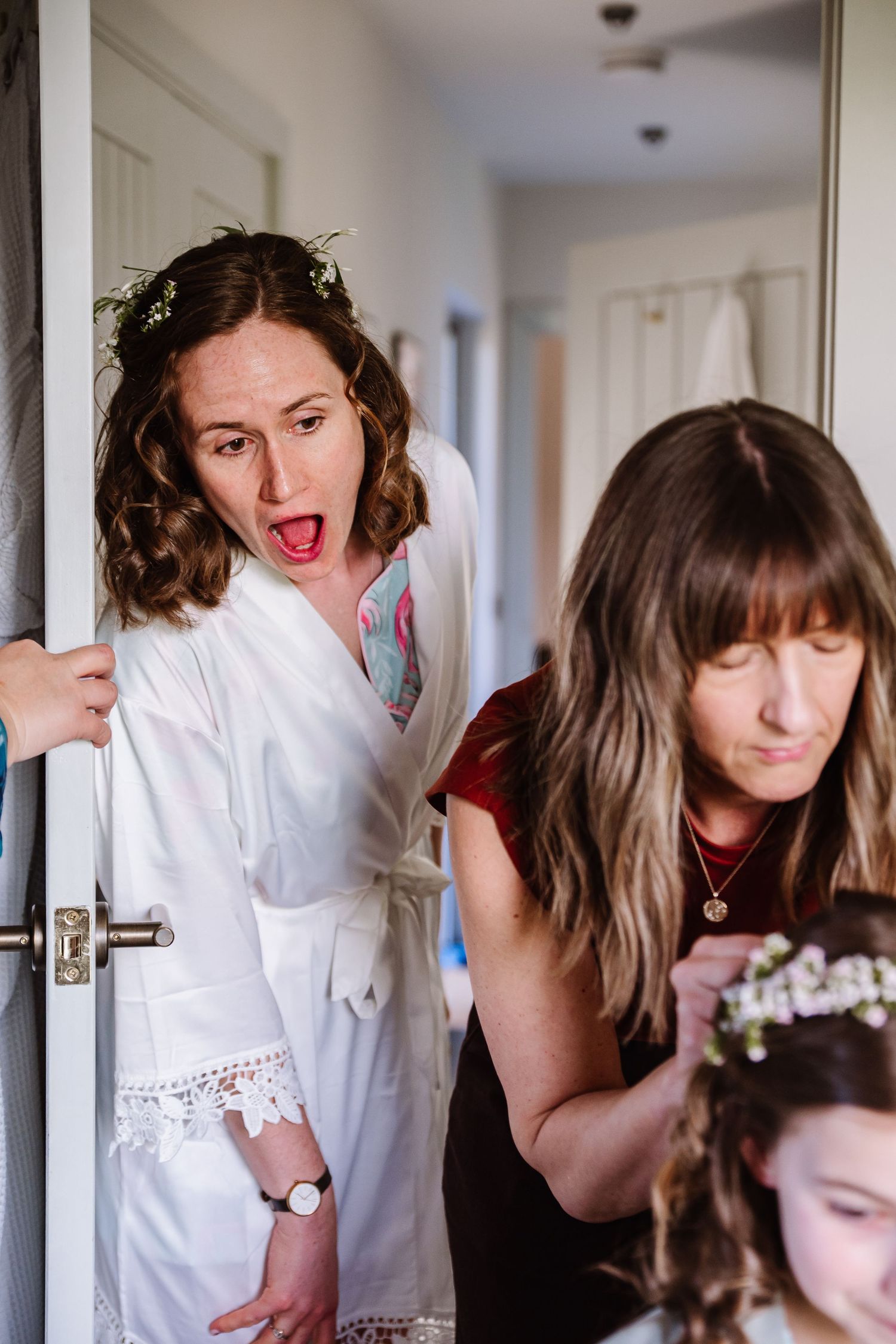 Bride in white robe and flower crown reacts with surprise as hairstylist arranges flowers in young girl's hair at The Cornish Place