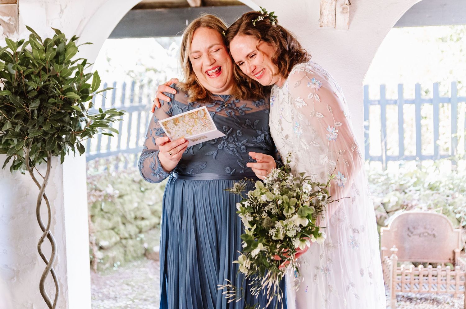 Bride and bridesmaid in blue gown sharing a joyful moment, looking at a card, with floral bouquet at outdoor wedding.