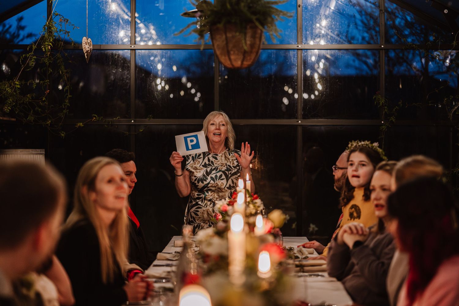 Woman holding a parking sign at a candlelit dinner party in a greenhouse with fairy lights and guests. The Cornish Place, Wood corni wedding