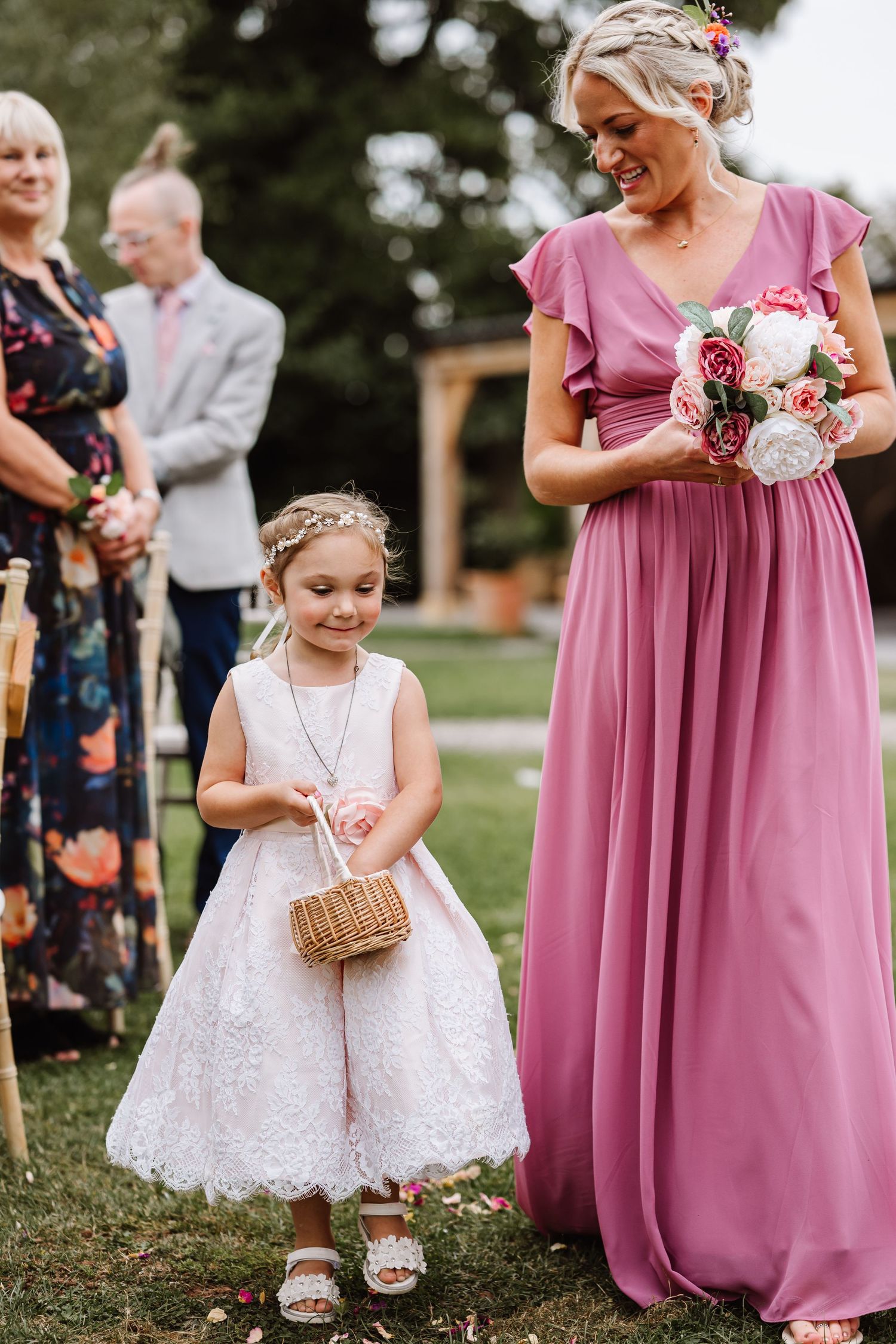 Little flower girls in white dresses and pink bridesmaids in mauve gowns gather outdoors during a wedding ceremony.
