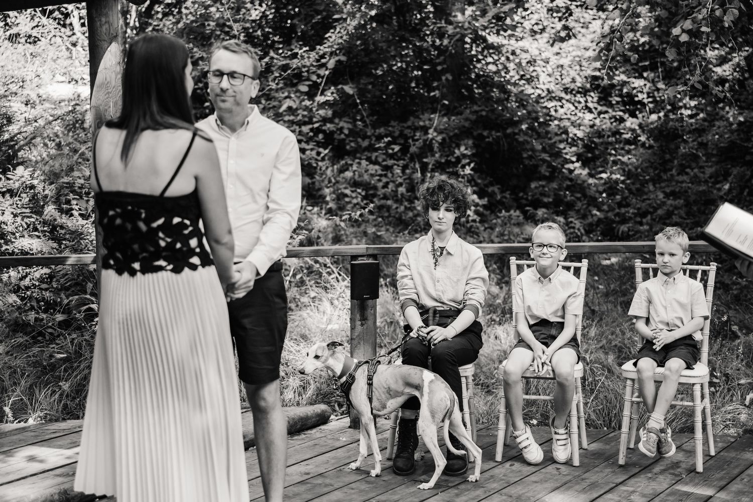 Moonacre, Dorset elopment Black and white outdoor wedding ceremony on a deck with guests seated, a dog present, surrounded by lush greenery.