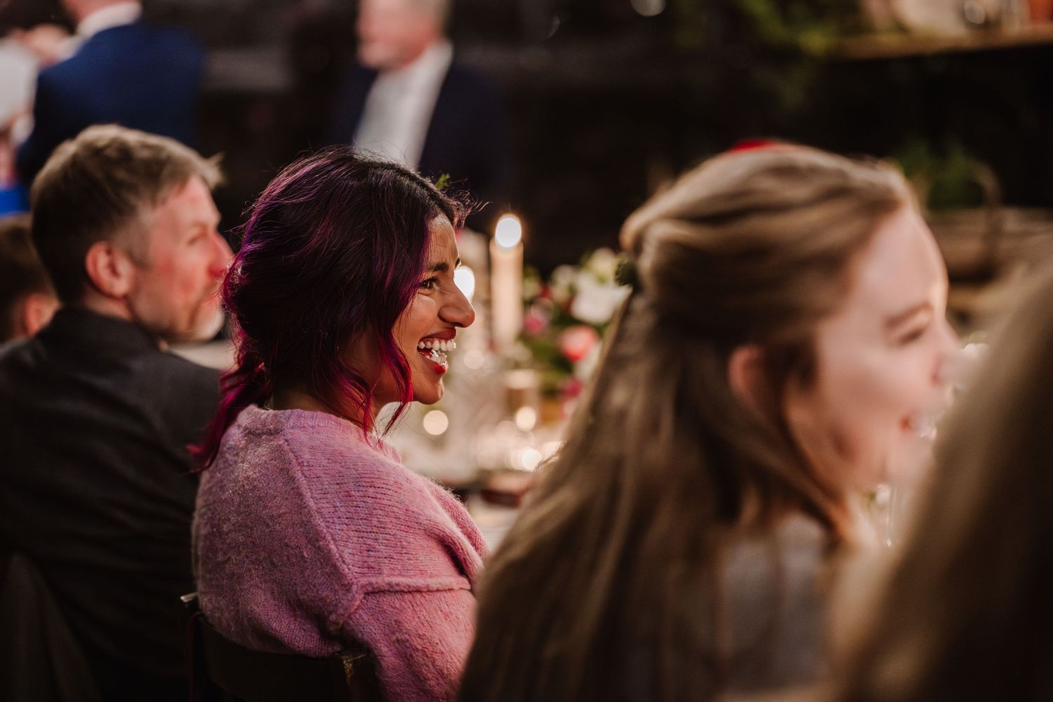 A woman with burgundy hair laughs joyfully at an elegant candlelit dinner event.