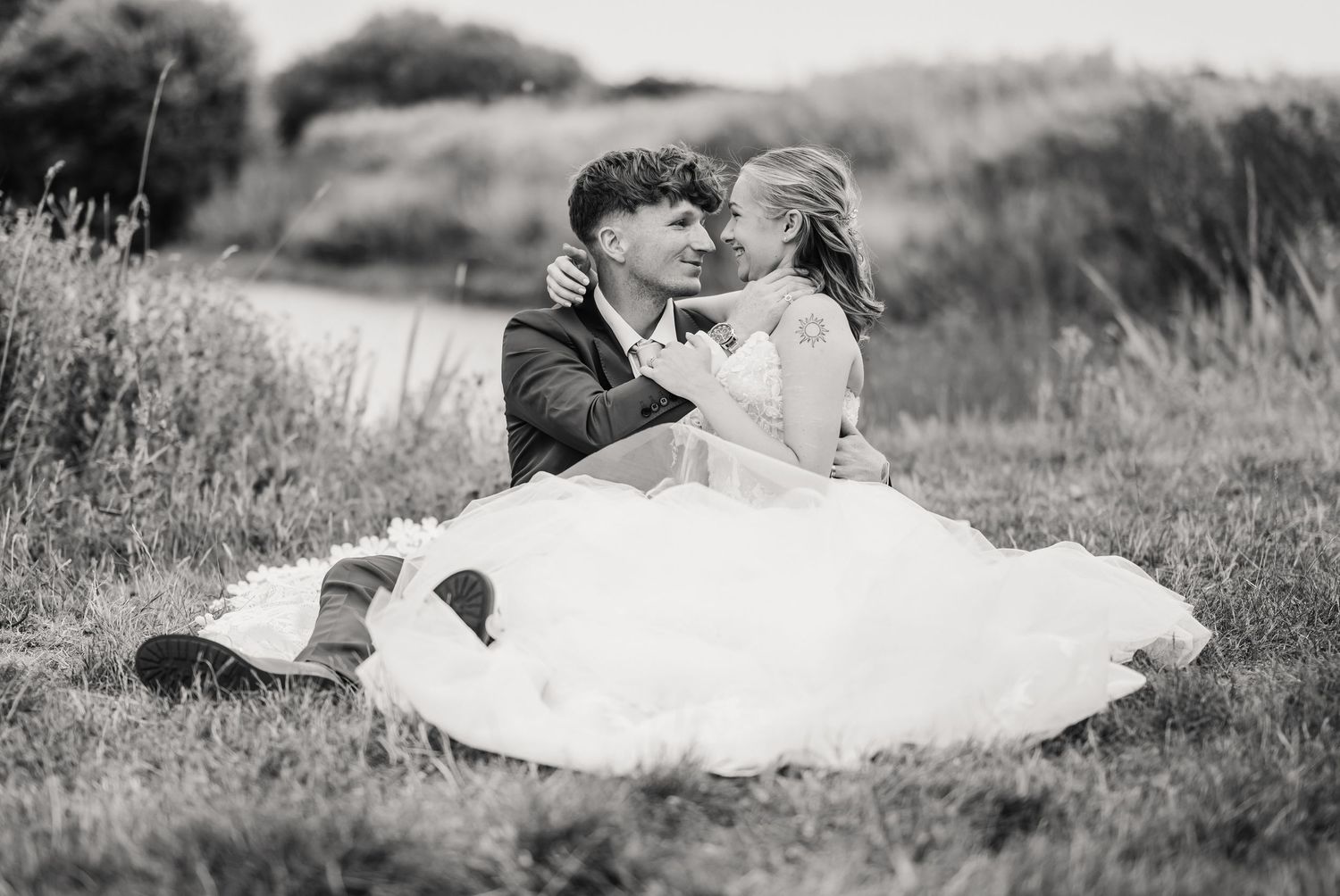 Black and white photo of a bride in a full tulle gown and a groom in a suit sitting close together in a grassy field, smiling and embracing.