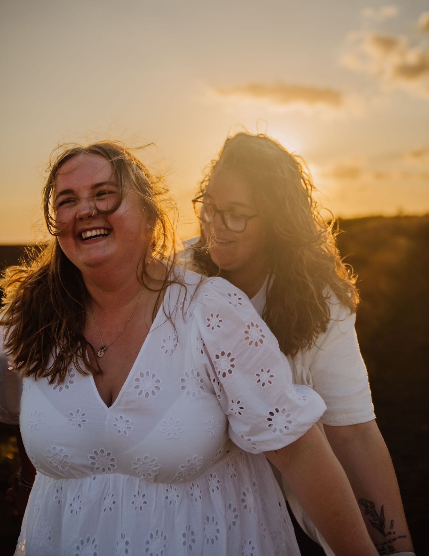 Two people in white dresses share joyful laughter together during golden hour sunset photoshoot. Relaxed Dorset Wedding photographer