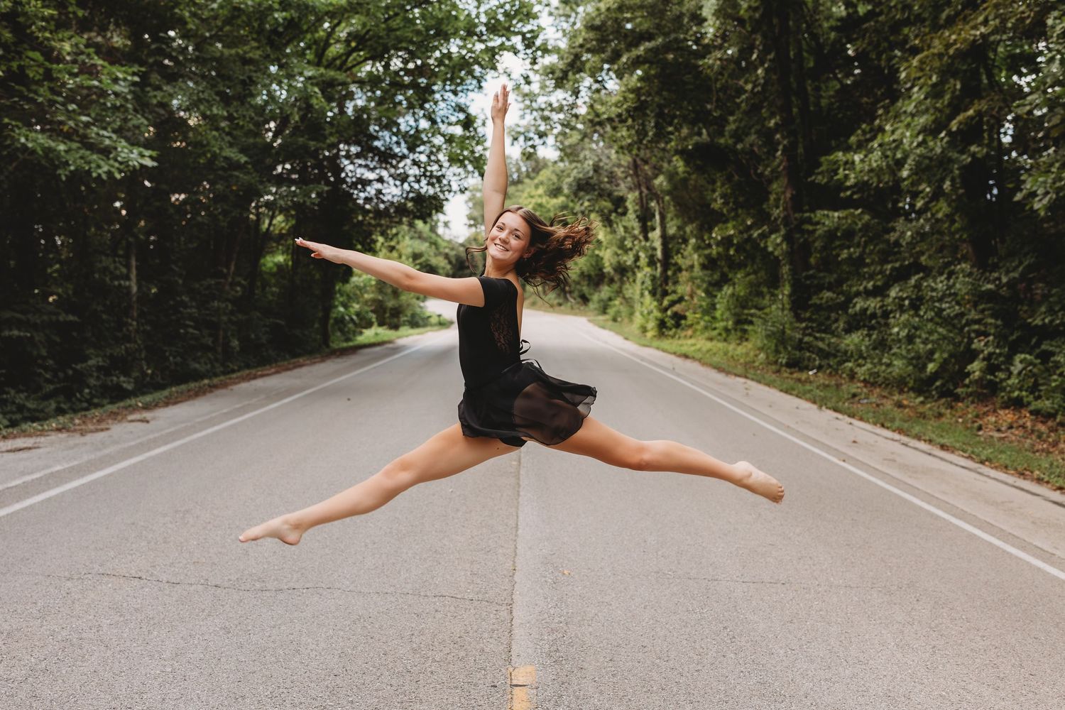 senior girl black leotard doing splits in the air in the middle of a road surrounded by trees