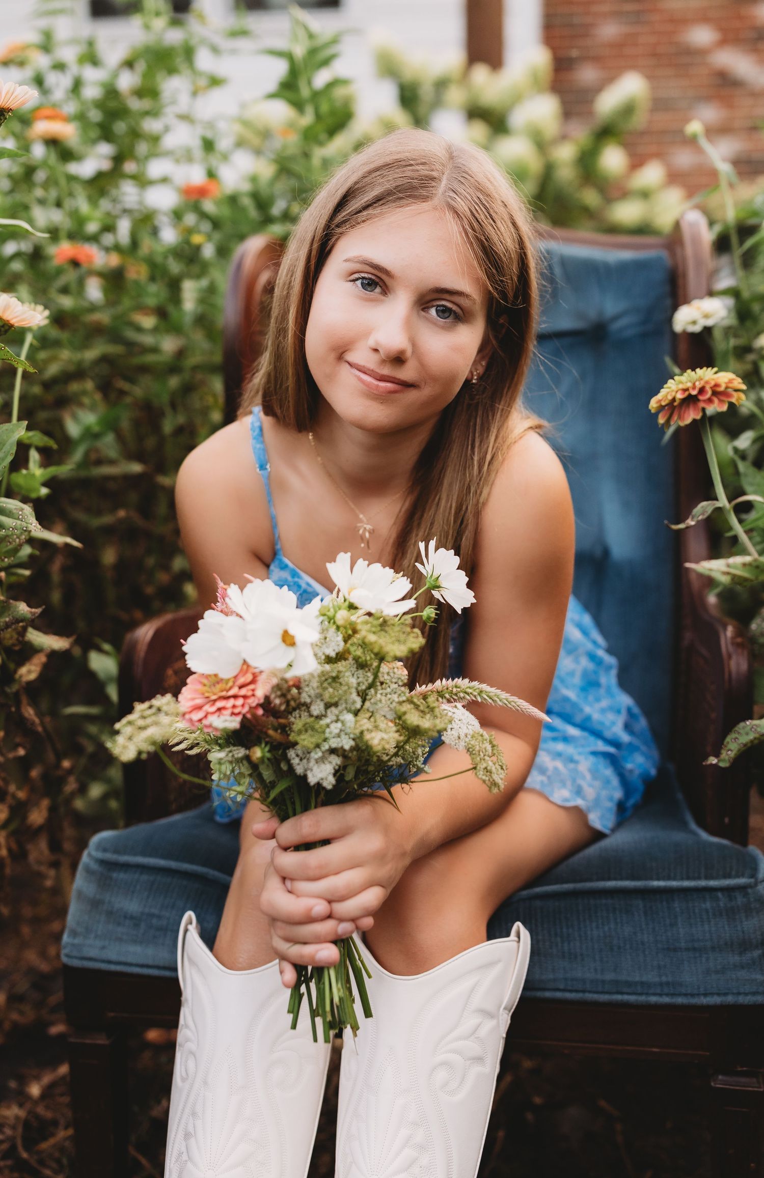 senior girl in the middle of a flower patch sitting in antique blue chair holding a bouquet of flowers white cowgirl boots and blue dress