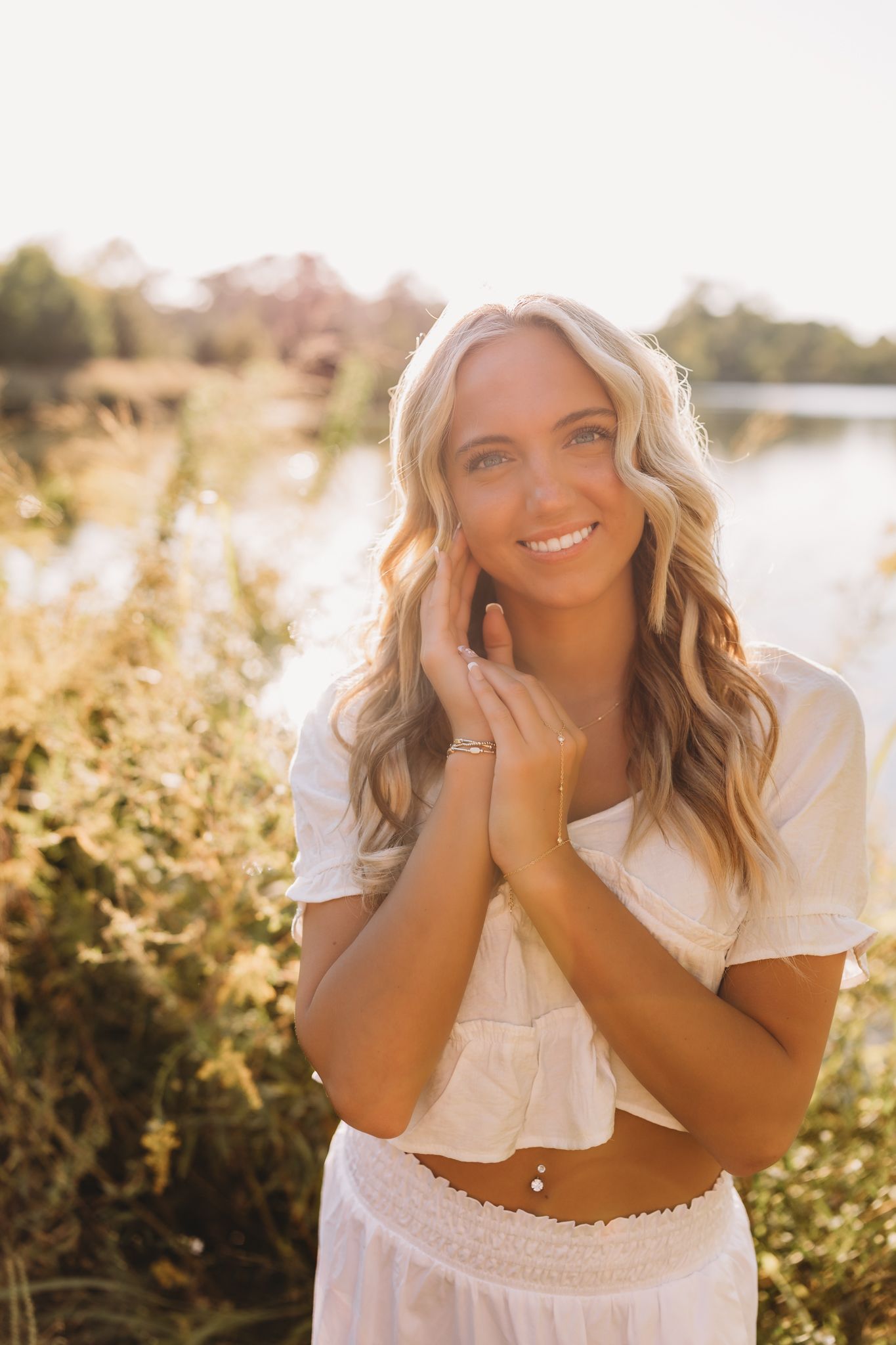 senior girl golden hour sun blonde hair blue eyes white shirt and skirt pond tall grass