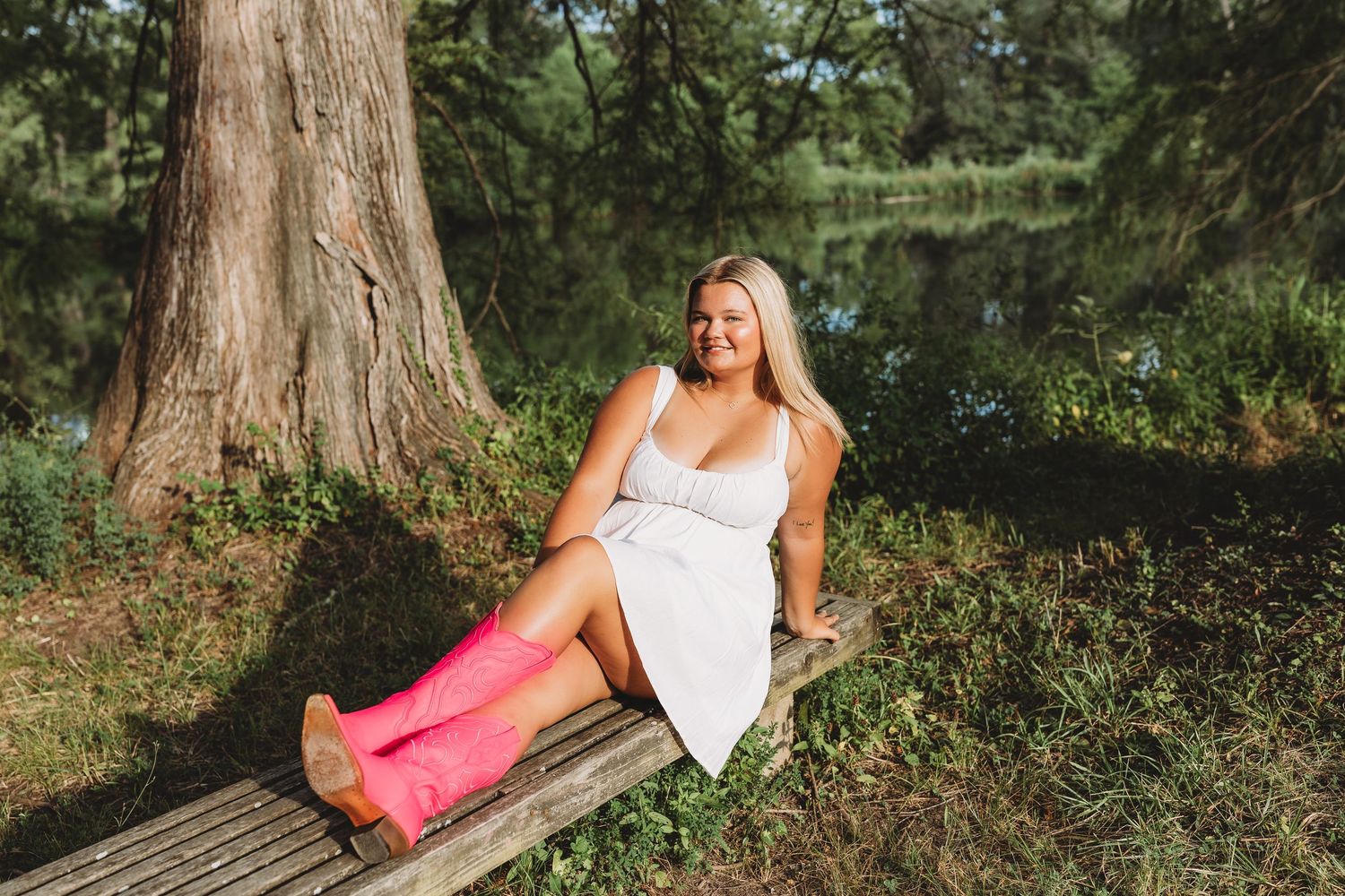 senior girl white dress hot pink cowgirl boots leaning back on a bench near a pond direct sunlight