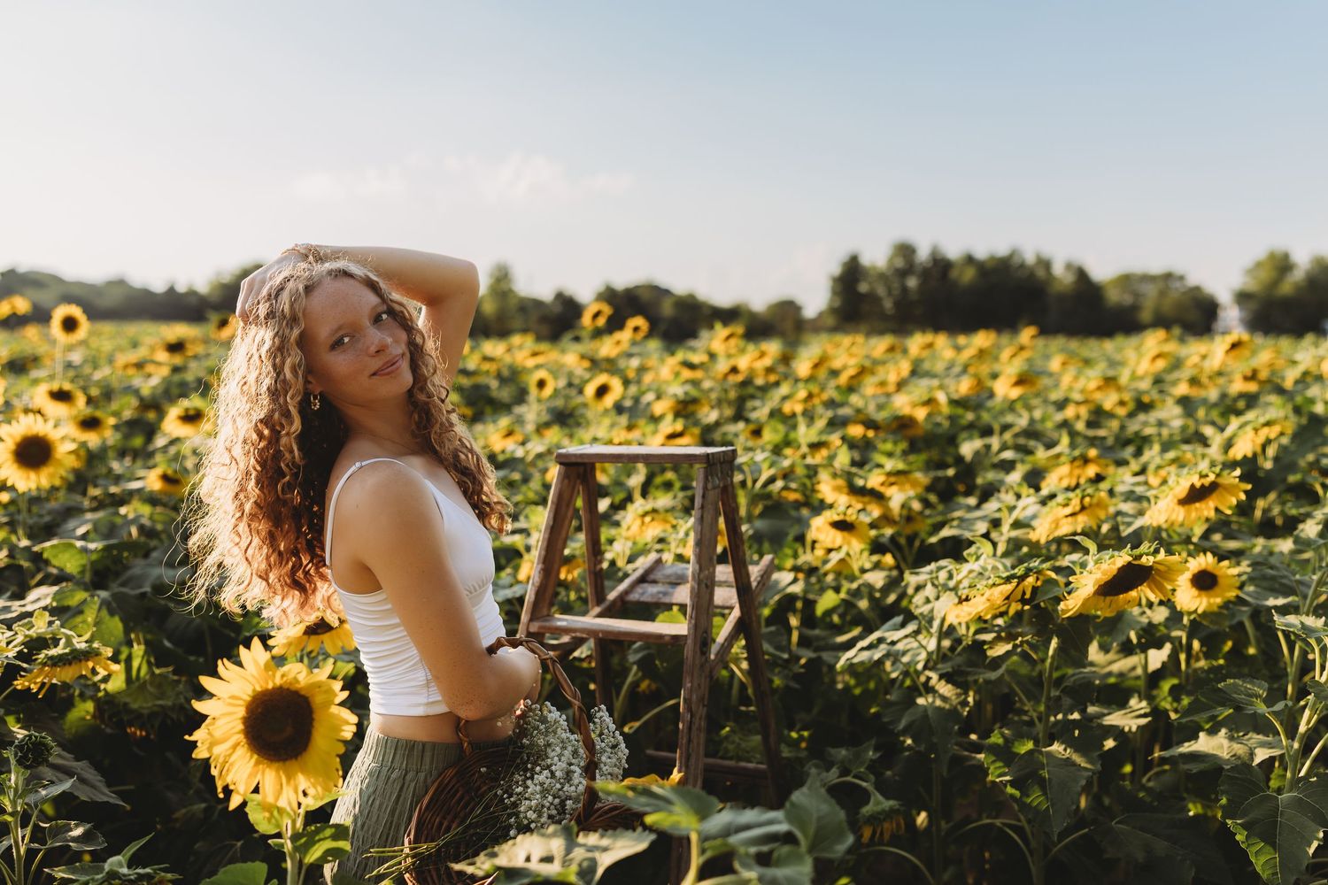 senior girl white top green dress in a field of sunflowers on a ladder