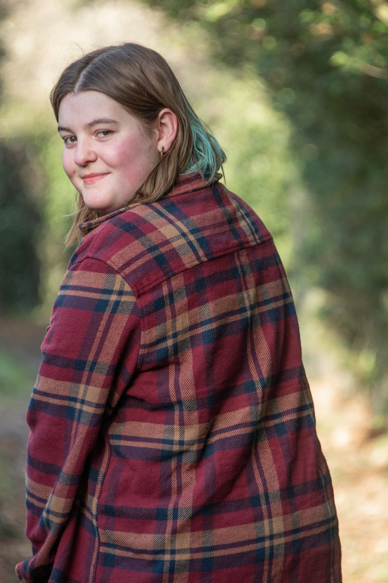 Young woman in red tartan flannel jacket glancing over shoulder on woodland path in natural light.