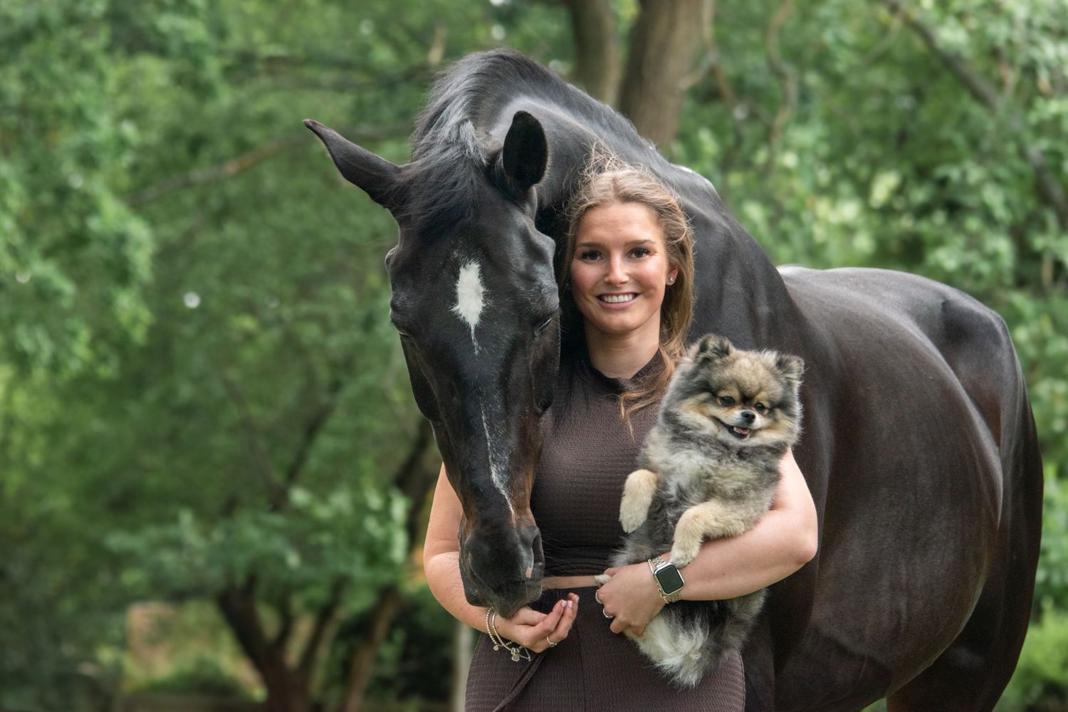 Blonde woman holding a Pomeranian dog standing beside a black horse in a lush garden.