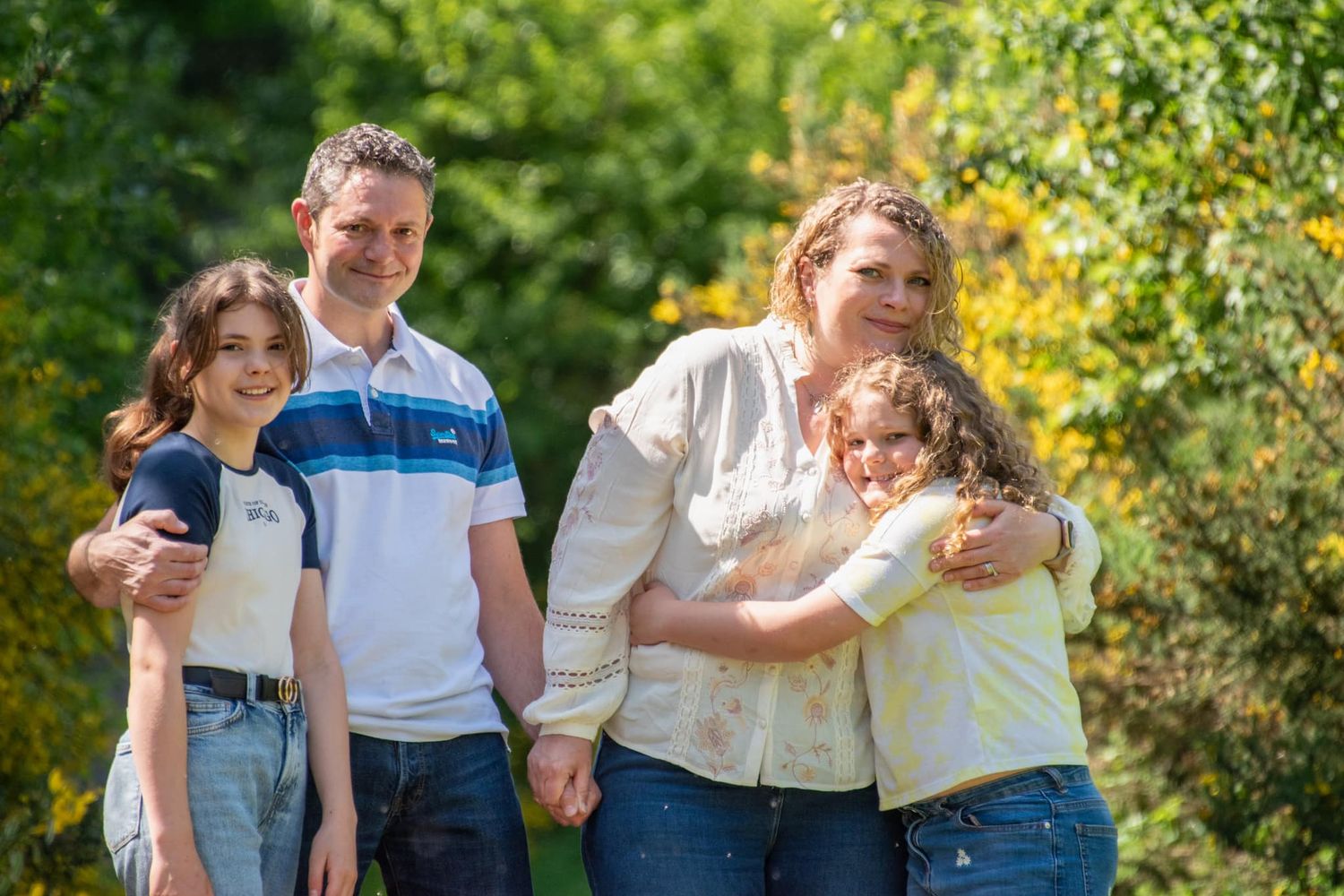 Happy family of four posing outdoors in a lush green park, parents holding children close while smiling.