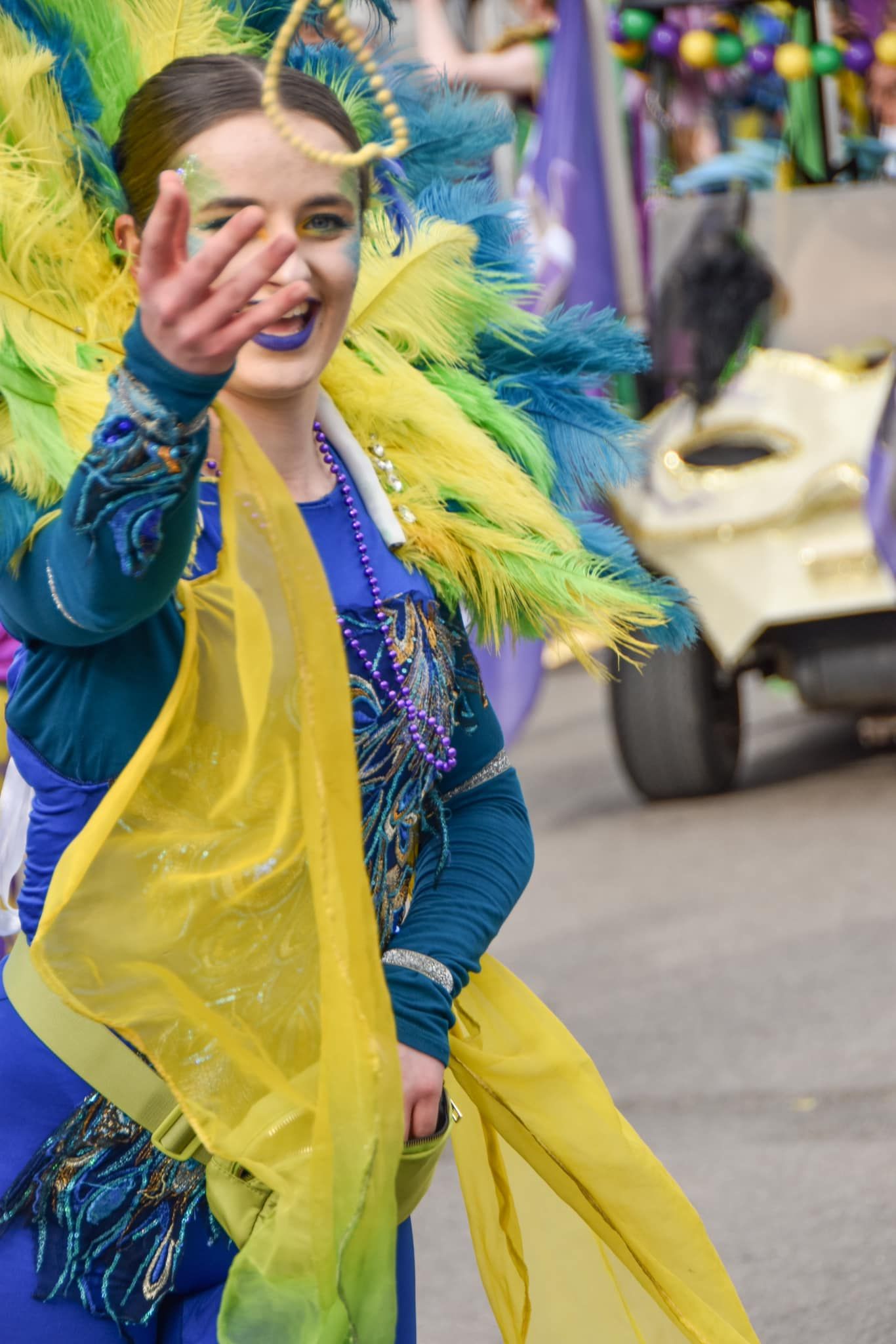 Mardi Gras parade performer in teal costume with yellow and green feathers waving to the crowd.