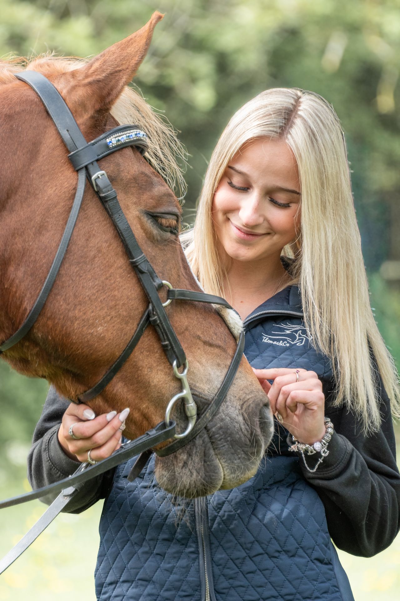 Young blonde woman smiling while feeding a chestnut horse outdoors.