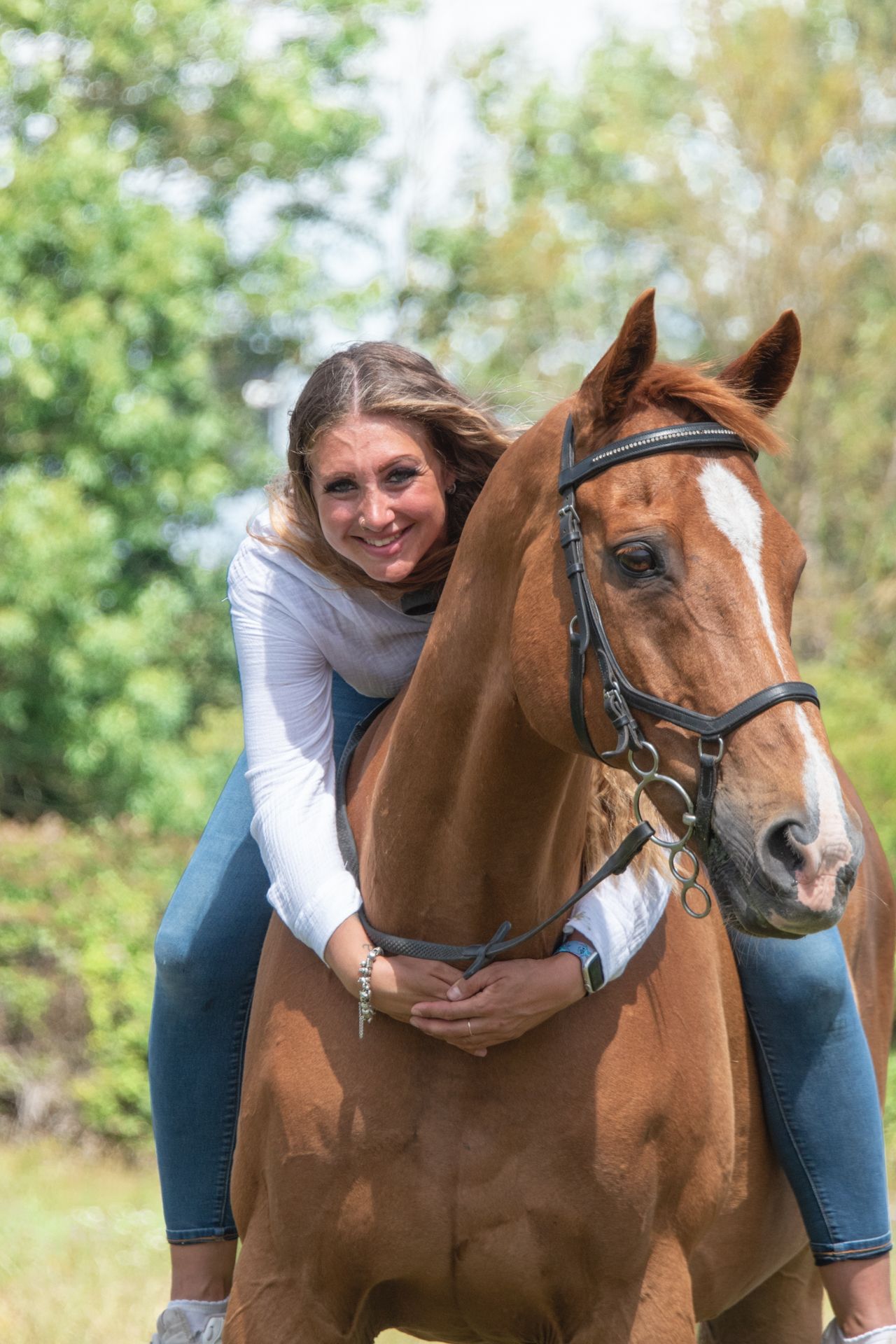 Smiling woman in white top hugging a chestnut horse with a blaze, outdoors among green trees.