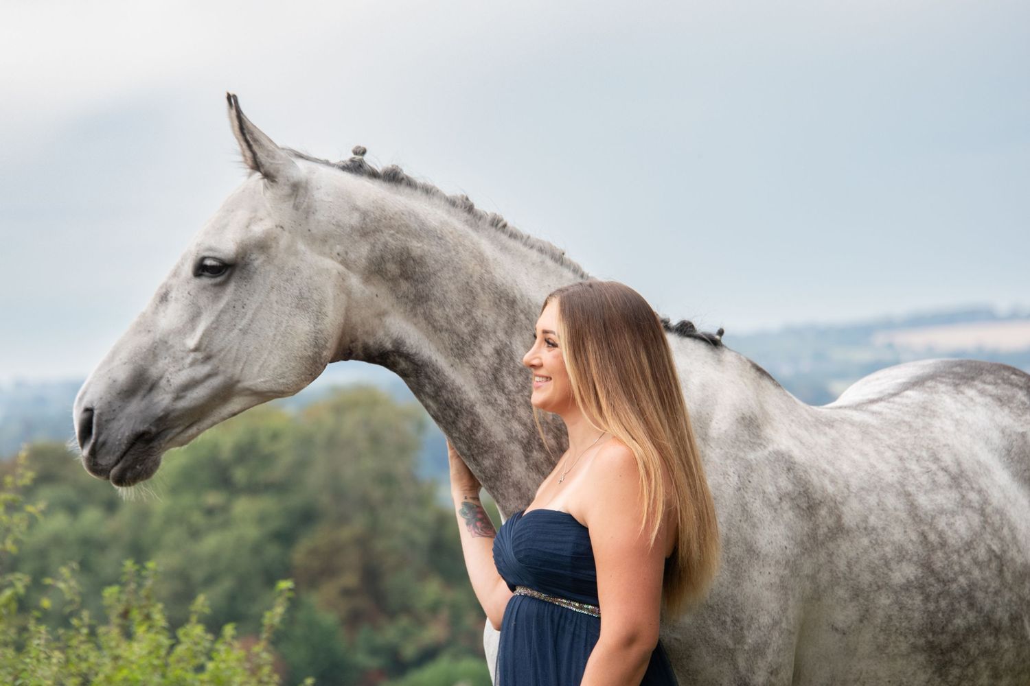A blonde woman in a navy dress stands beside a dapple grey horse in an outdoor setting.