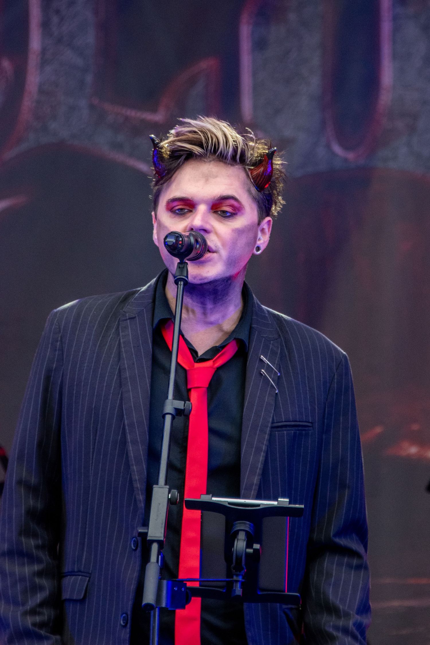 Male singer in pinstripe suit with devil horns makeup performing at microphone on dark stage.