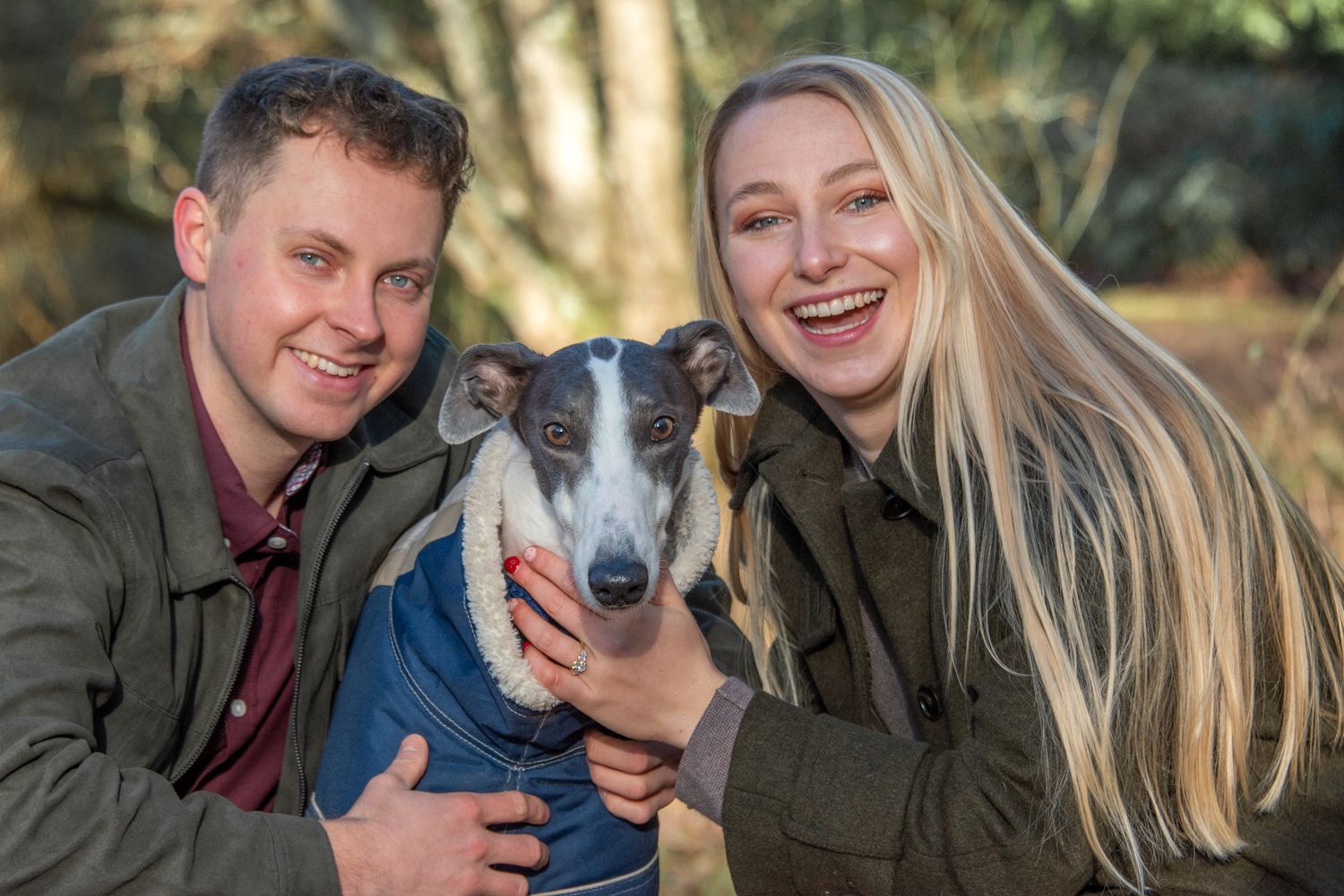 Happy engaged couple smiling outdoors with their grey and white whippet dog wearing a blue coat.